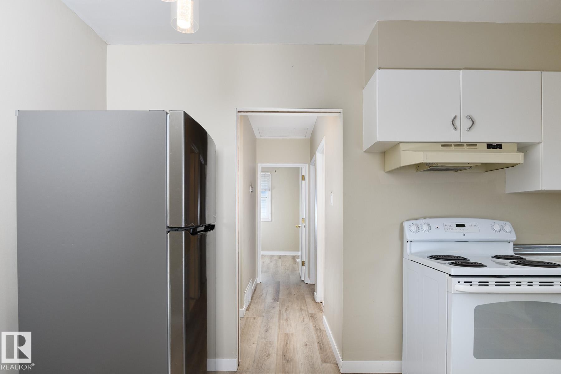 Kitchen area featuring a stainless steel refrigerator, white electric range, and white upper cabinetry - 9607 75 Street, Edmonton, AB