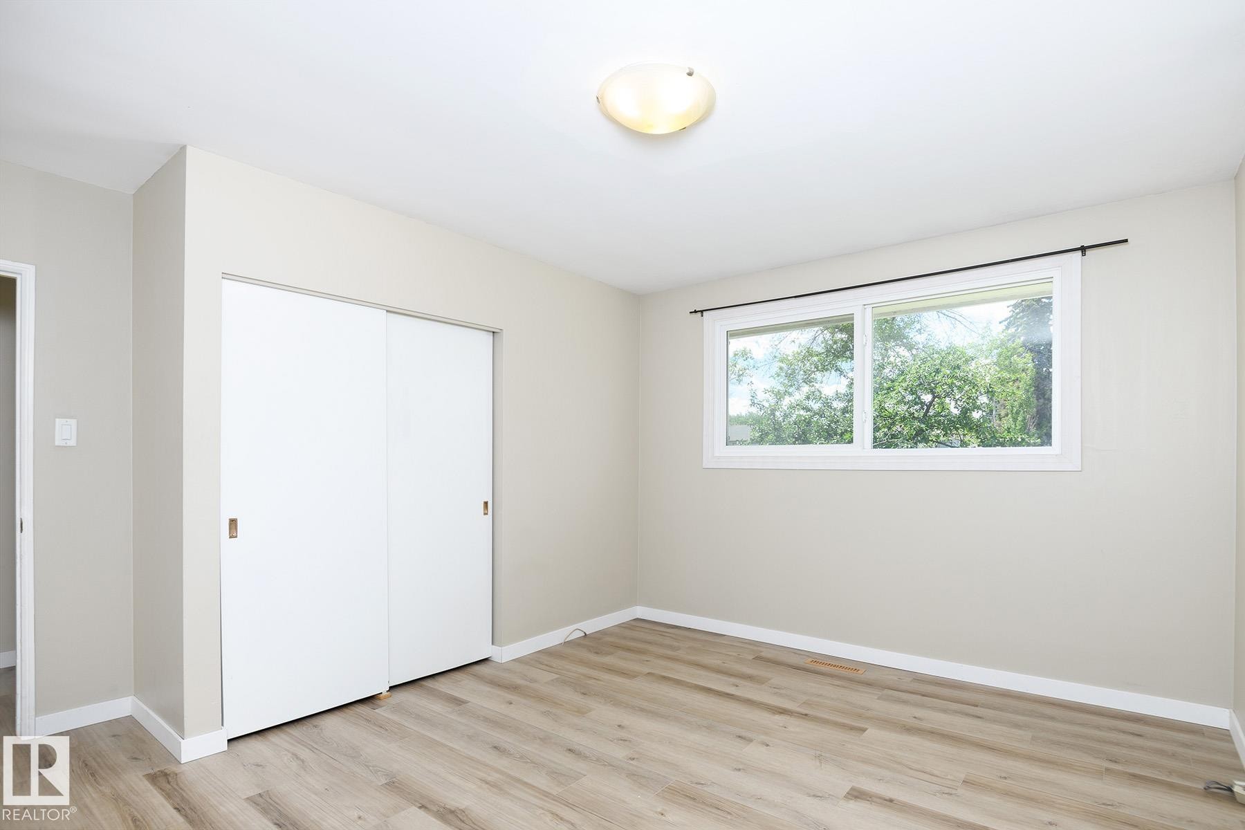 Interior room featuring a large horizontal window, wood-finish flooring, white baseboards, and a built-in closet with sliding doors - 9607 75 Street, Edmonton, AB