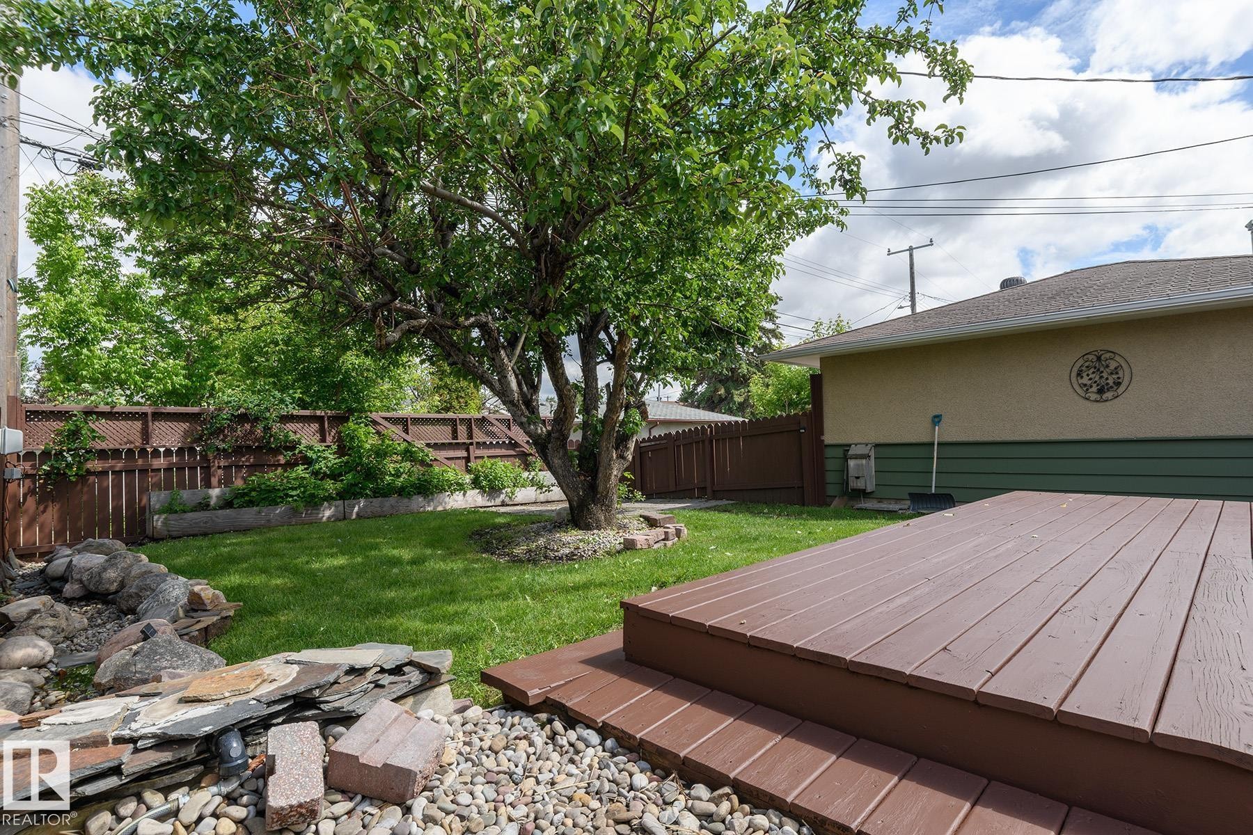 Expansive backyard featuring a large mature tree, a brown privacy fence with lattice accents, a multi-level wood-finish deck, and a rock garden with varied stone elements - 9607 75 Street, Edmonton, AB