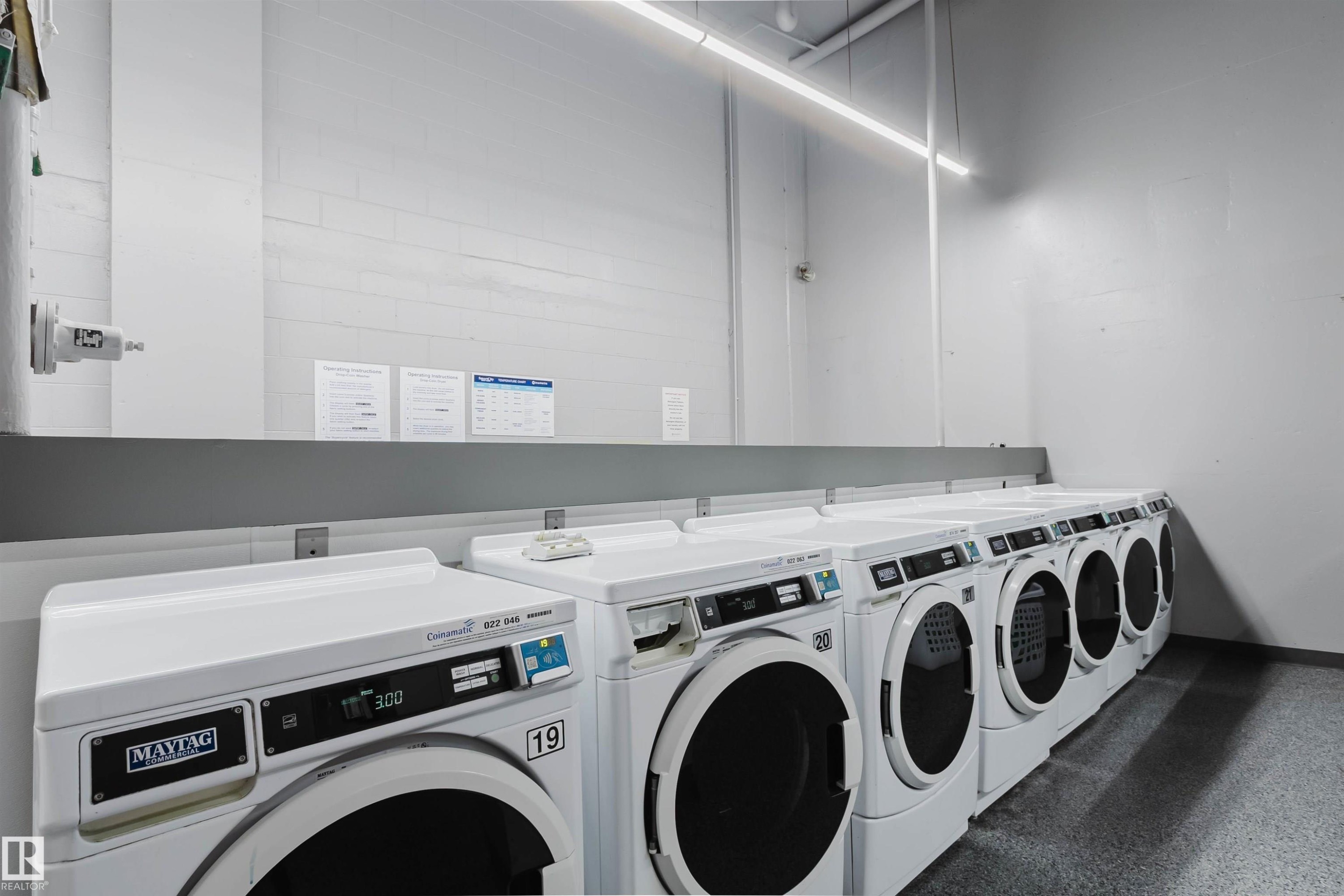 On-site laundry facility featuring multiple front-loading washers and dryers, white appliances, durable flooring, and ample overhead lighting - 904 10883 Saskatchewan Drive, Edmonton, AB