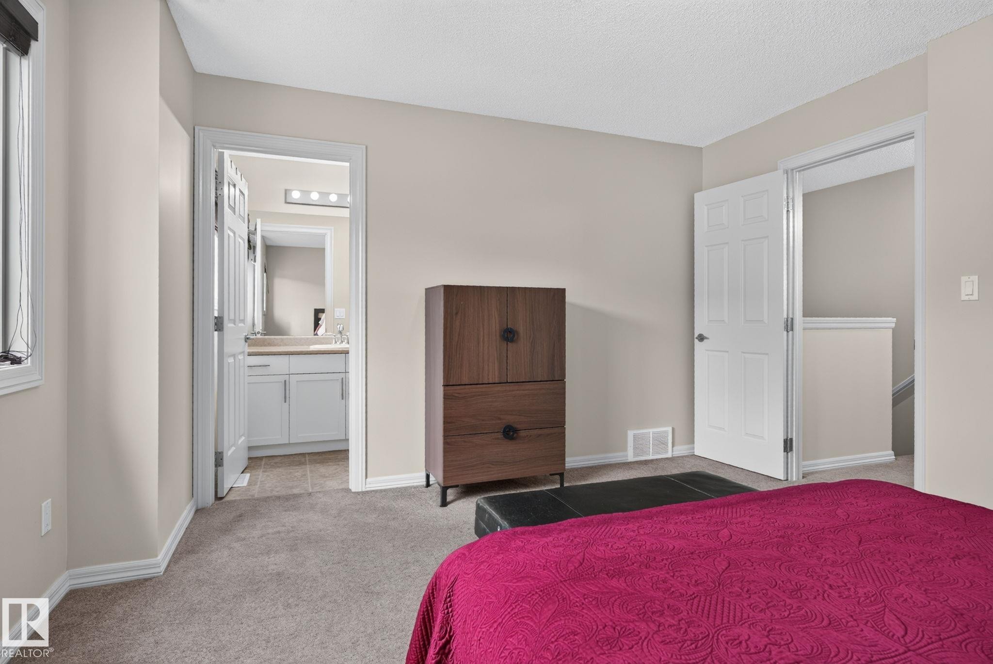 Carpeted room with light neutral wall paint and white trim, featuring a window with blinds, a white paneled door, and a second doorway revealing a bathroom with a vanity and mirror - 545 Watt Boulevard Sw, Edmonton, AB - Indoor Photo Showing Bedroom
