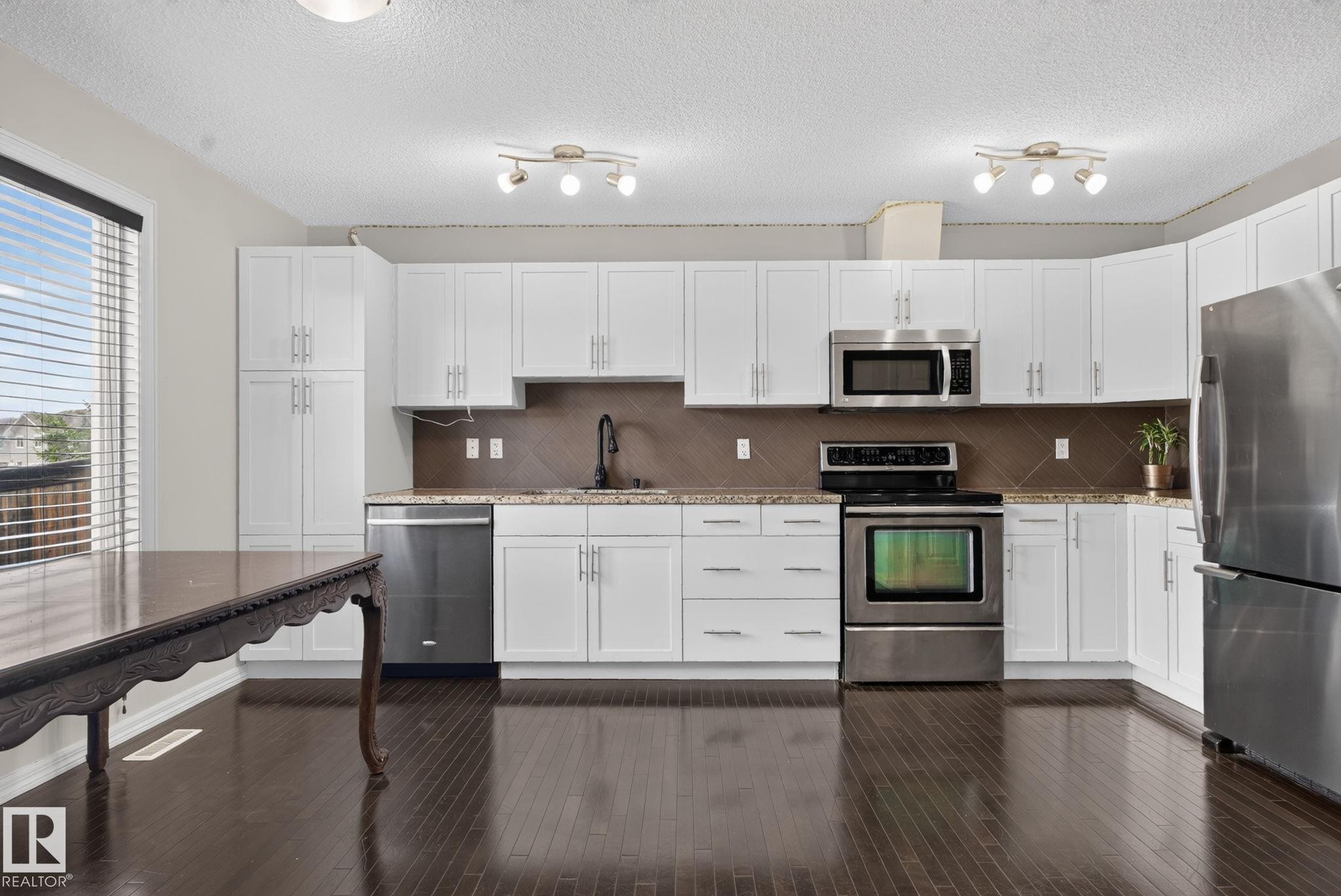 Contemporary kitchen featuring white cabinetry, stainless steel appliances, dark wood-finish flooring, granite countertops, and a tiled backsplash - 545 Watt Boulevard Sw, Edmonton, AB - Indoor Photo Showing Kitchen With Stainless Steel Kitchen With Upgraded Kitchen