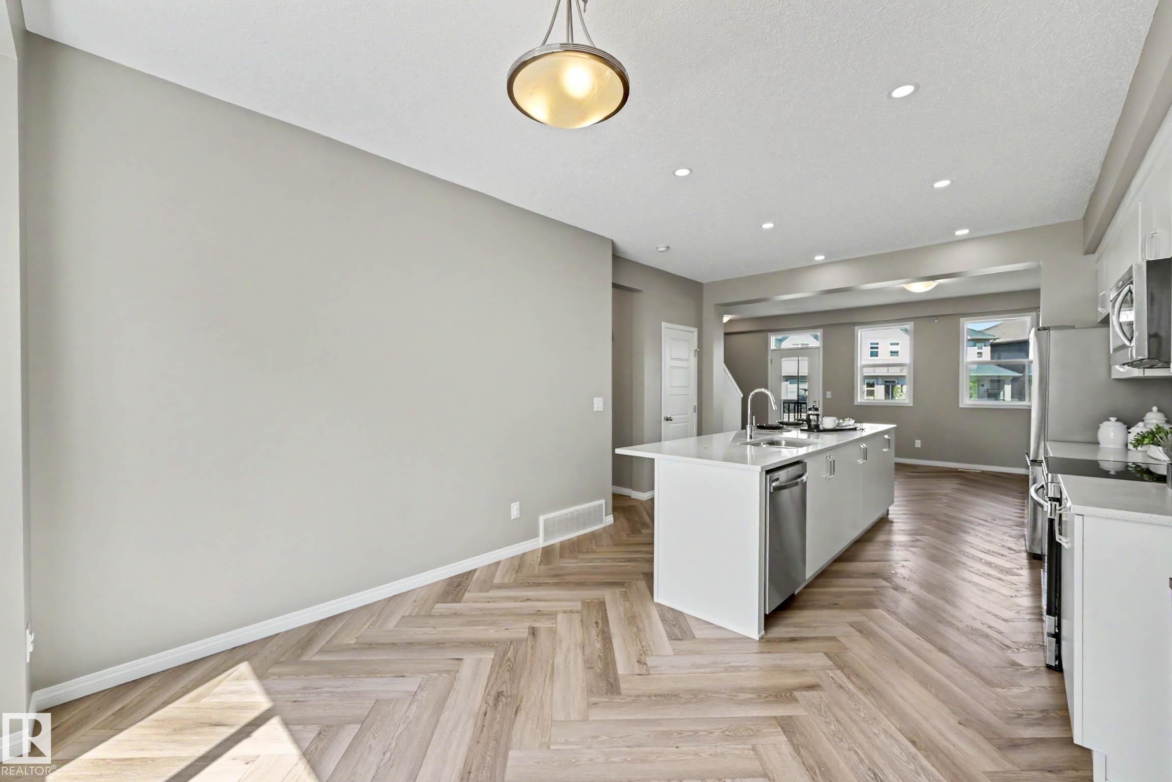 Open-concept living space featuring wood-finish flooring in a herringbone pattern, a central kitchen island with an undermount sink, recessed lighting, and stainless steel appliances - 53 2072 Wonnacott Way, Edmonton, AB
