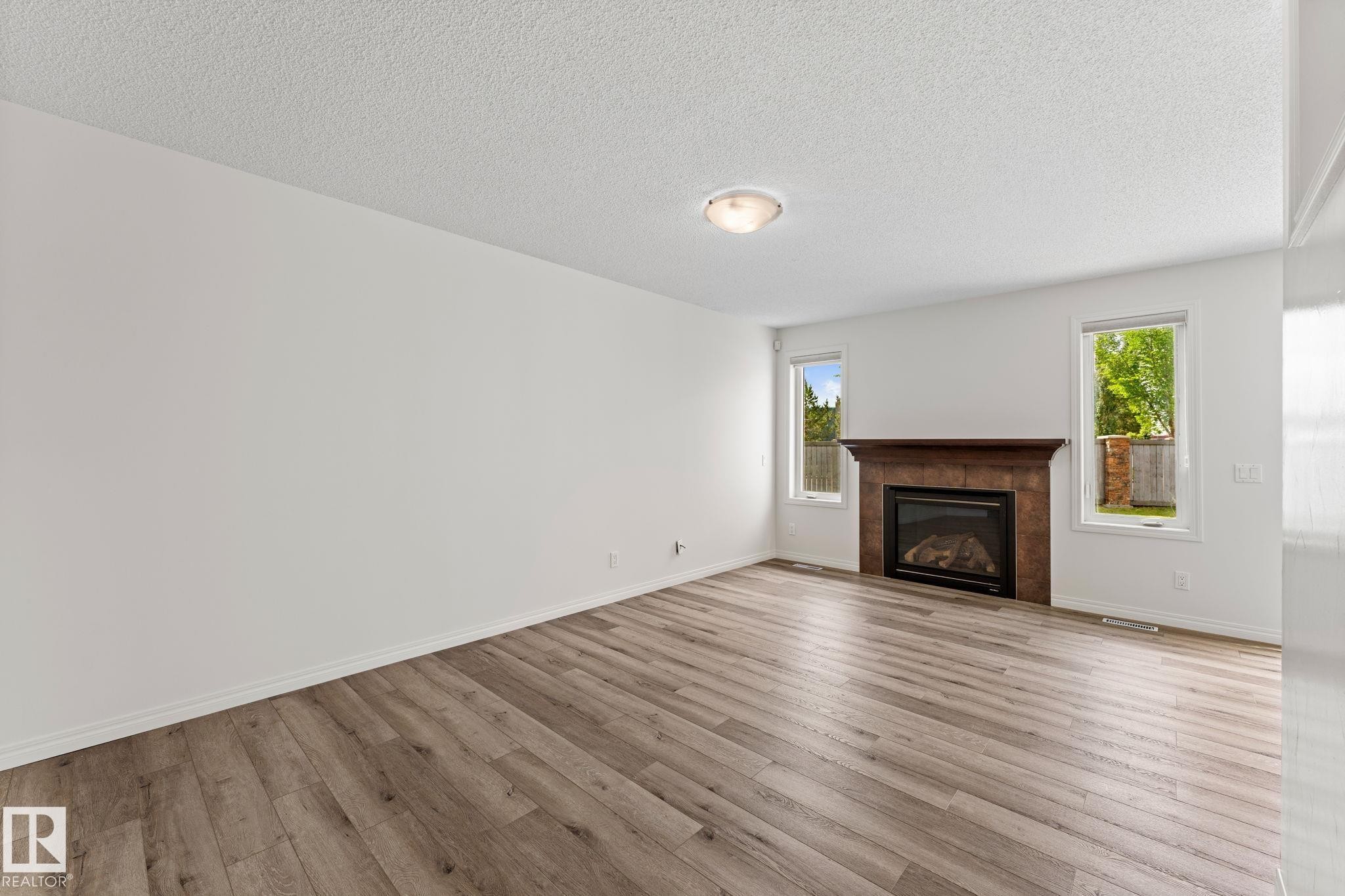 Spacious room featuring wood-finish flooring, a contemporary fireplace with a stone surround and wood mantel, white walls, and two windows providing natural light - 12523 171 Avenue, Edmonton, AB