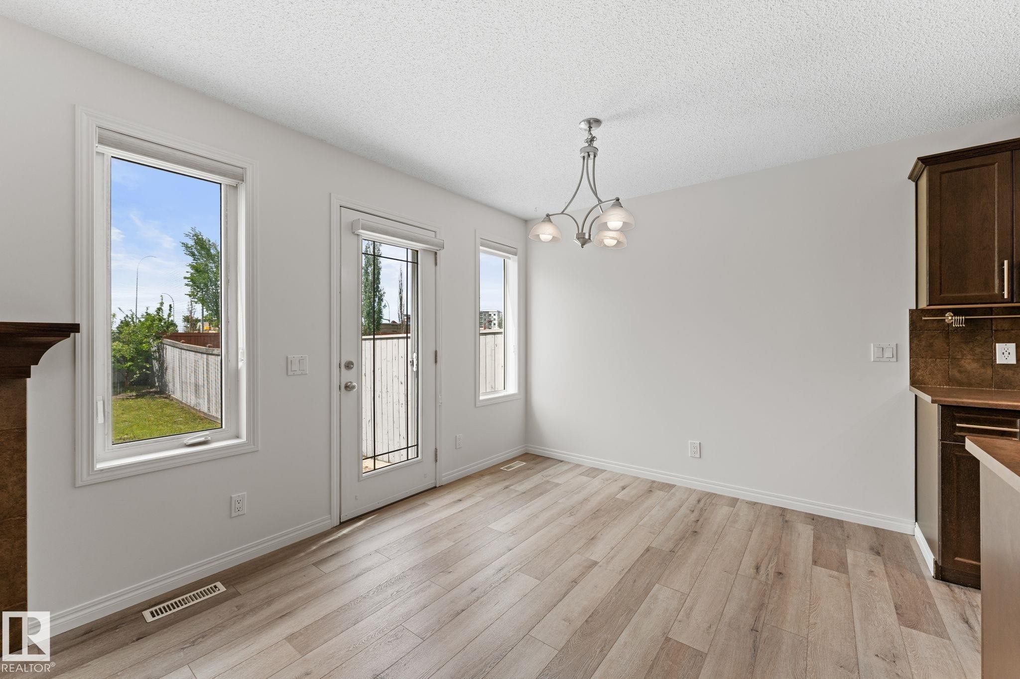 Spacious dining area featuring wood-finish flooring, ample natural light from multiple windows, a glass-paned door, a contemporary chandelier, and rich wood cabinetry with a coordinating backsplash - 12523 171 Avenue, Edmonton, AB