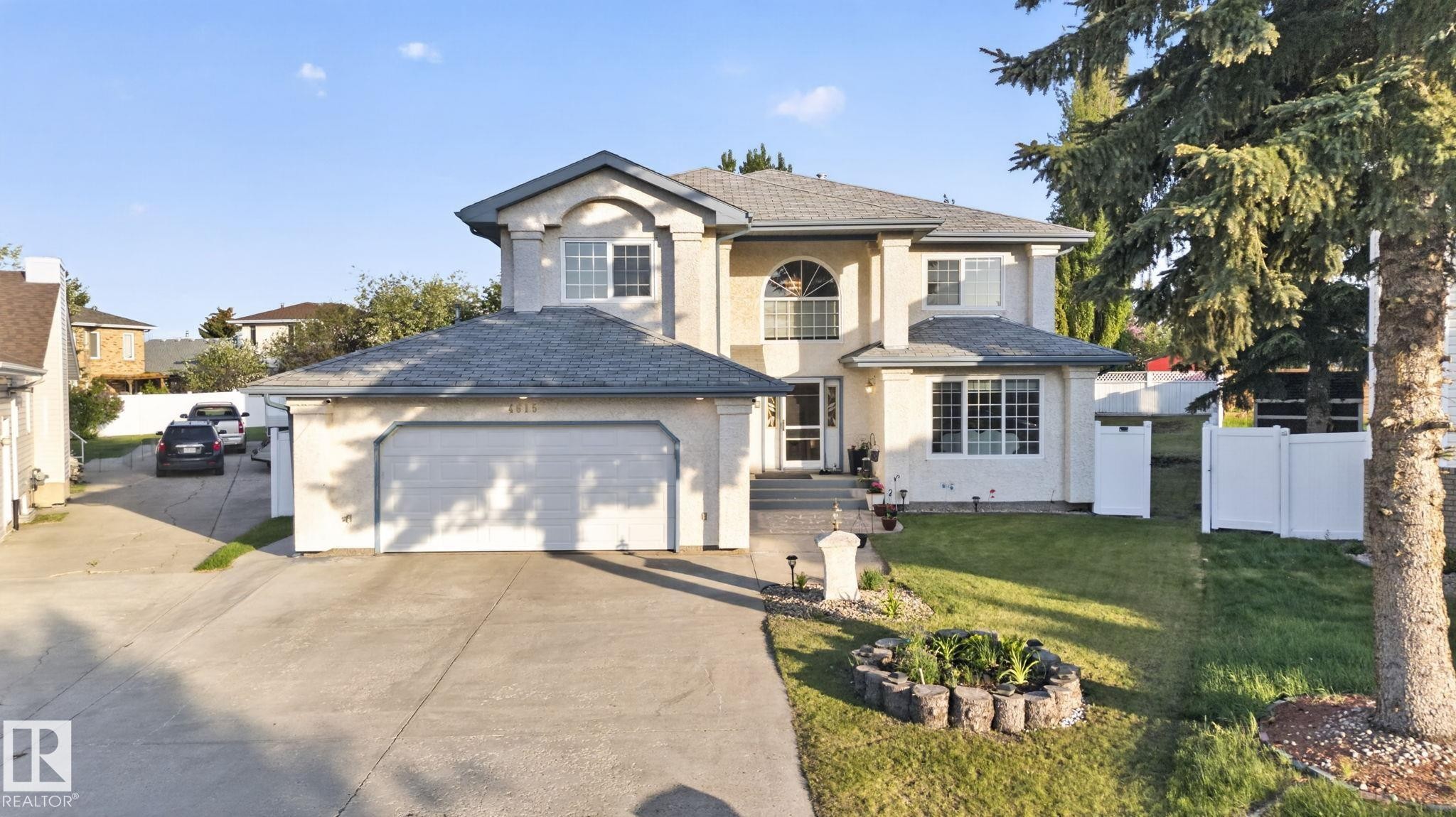 Stucco exterior residence featuring a two-car attached garage, arched window accents, a front lawn with established landscaping, and a side yard with white fencing - 4615 43A Avenue, Edmonton, AB - Outdoor