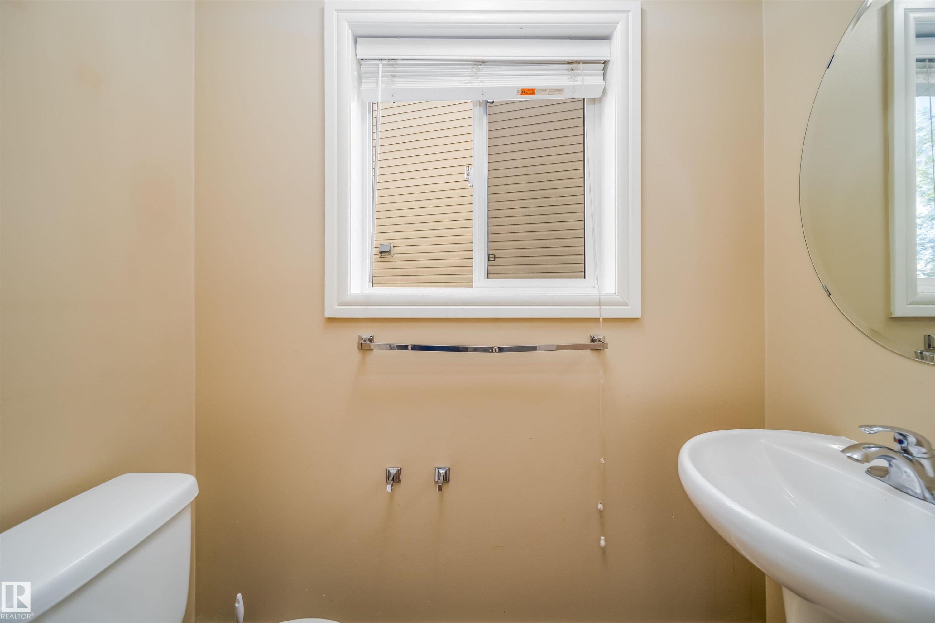 Compact bathroom featuring a white porcelain toilet, a pedestal sink with a chrome faucet, a round wall-mounted mirror, and a window with white trim - #46 15151 43 St Nw, Edmonton, AB - Indoor Photo Showing Bathroom
