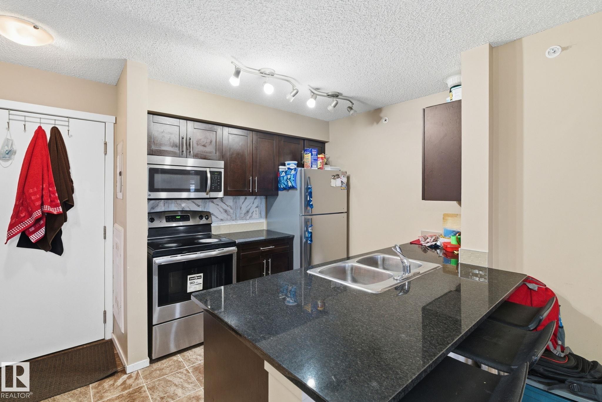 Kitchen featuring a dual-basin stainless steel sink, dark wood-finish cabinetry, stainless steel appliances, speckled dark countertops, and a track lighting fixture - 424 1510 Watt Drive, Edmonton, AB