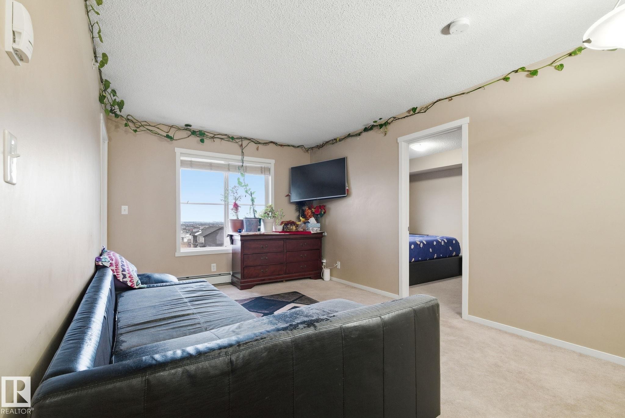 Carpeted interior space featuring a window with exterior visibility, light beige wall paint, a white ceiling, and a doorway opening to an additional room - 424 1510 Watt Drive, Edmonton, AB