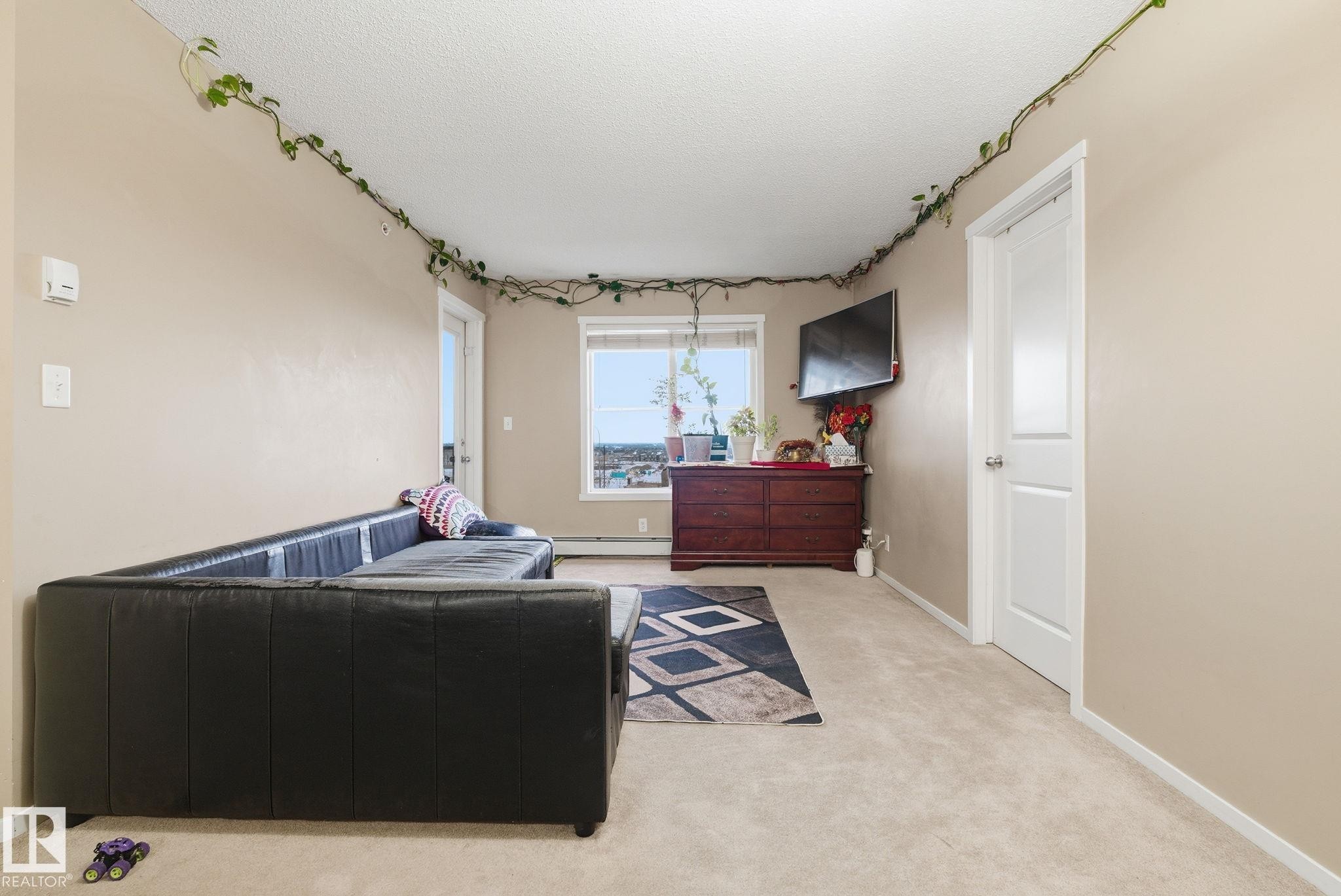 Carpeted room featuring a window with a cityscape backdrop, a built-in wall-mounted television, and a white interior door - 424 1510 Watt Drive, Edmonton, AB