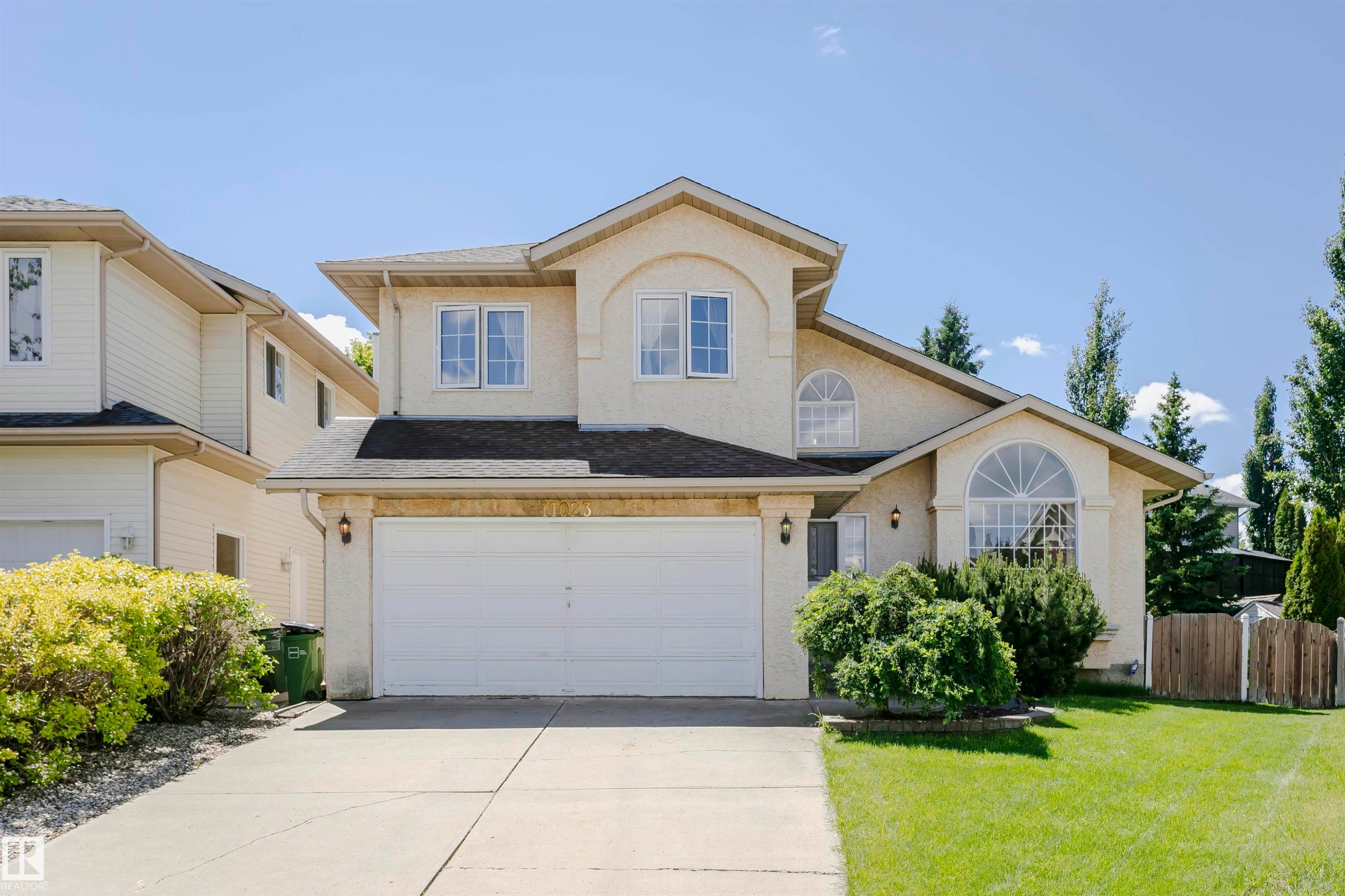Stucco exterior featuring a dual-car garage, concrete driveway, arched window, and mature landscaping - 11023 11A Avenue Nw, Edmonton, AB - Outdoor