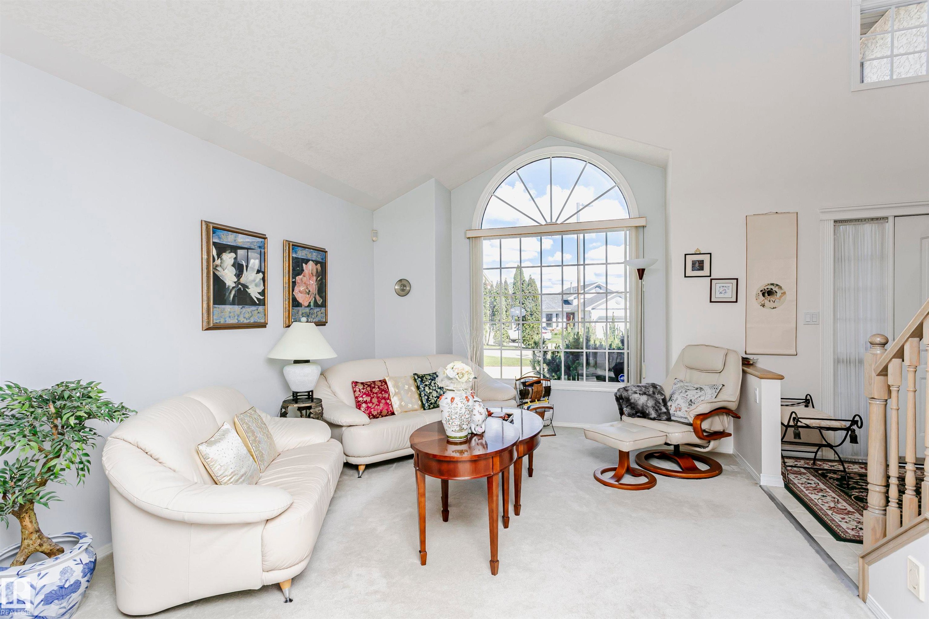 Vaulted ceiling living area featuring a large arched window, light-colored carpeting, a partial staircase with wood railing, and a windowed entryway - 11023 11A Avenue Nw, Edmonton, AB - Indoor Photo Showing Living Room