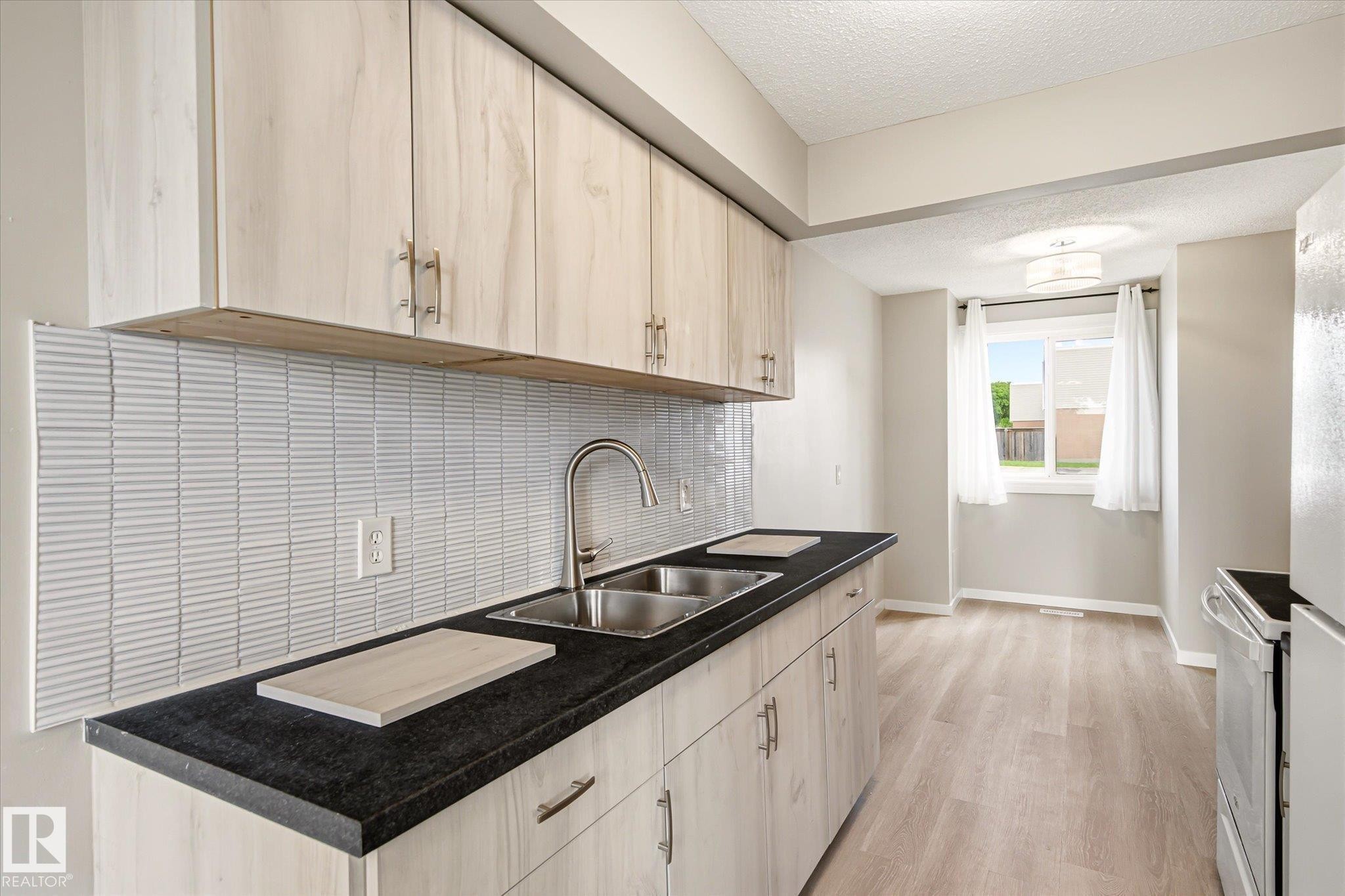 Kitchen featuring light wood-finish cabinetry with silver hardware, dark countertops, and a stainless steel double sink - 186 Cornell Crest, Edmonton, AB