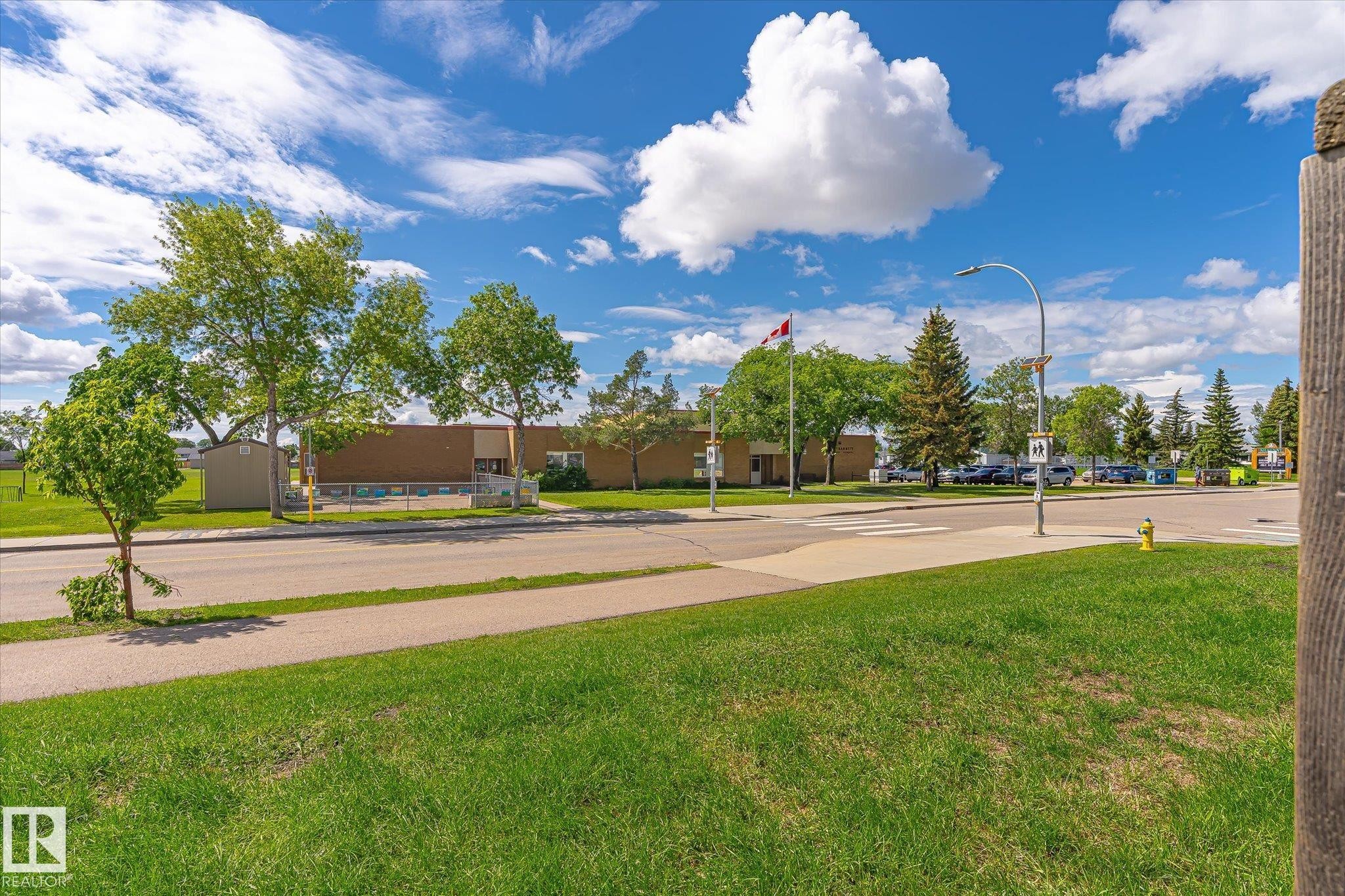 Grassy expanse with paved sidewalk and street, featuring mature trees and a distant building with a Canadian flag - 186 Cornell Crest, Edmonton, AB