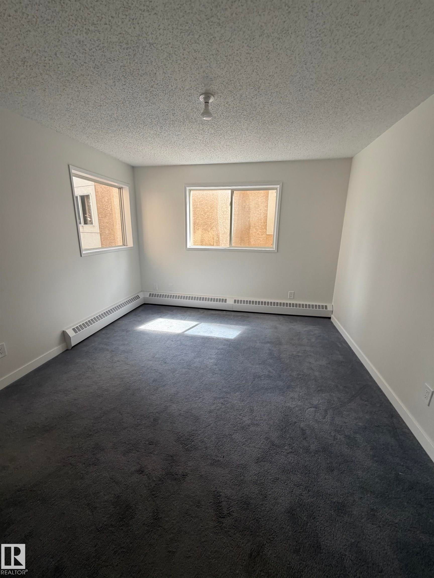 Carpeted room featuring two windows, white walls, and a popcorn ceiling - 213 17467 98A Avenue, Edmonton, AB