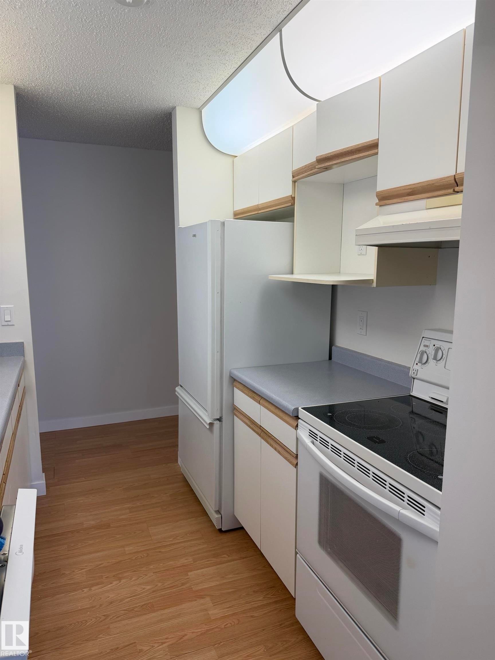 Galley kitchen featuring wood-finish flooring, white cabinetry with wood trim accents, light-toned countertops, and a full suite of white appliances including a refrigerator and electric range - 213 17467 98A Avenue, Edmonton, AB