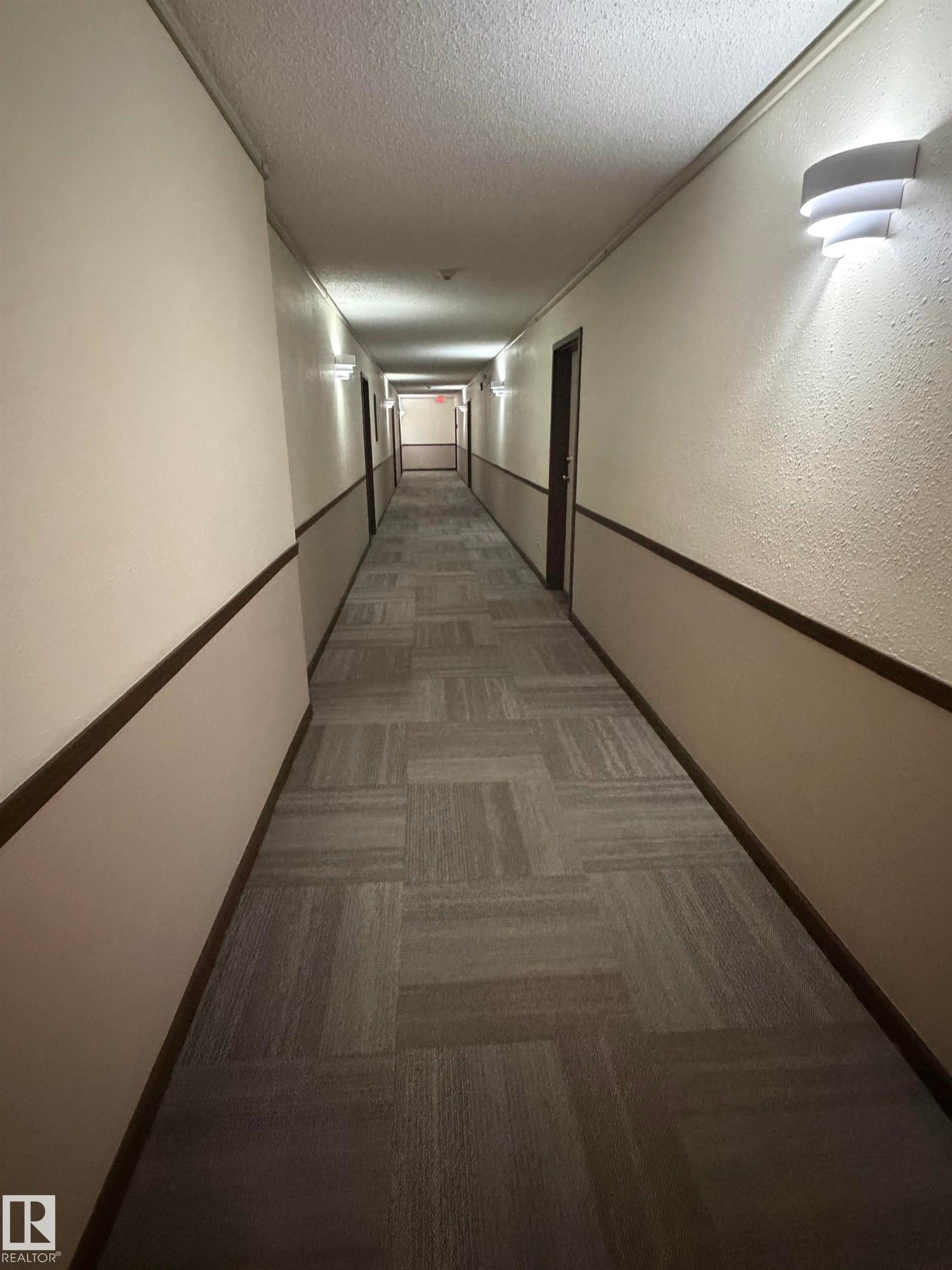 Long hallway featuring patterned carpet flooring, two-tone wall treatment, dark wood trim, and wall-mounted sconce lighting - 213 17467 98A Avenue, Edmonton, AB