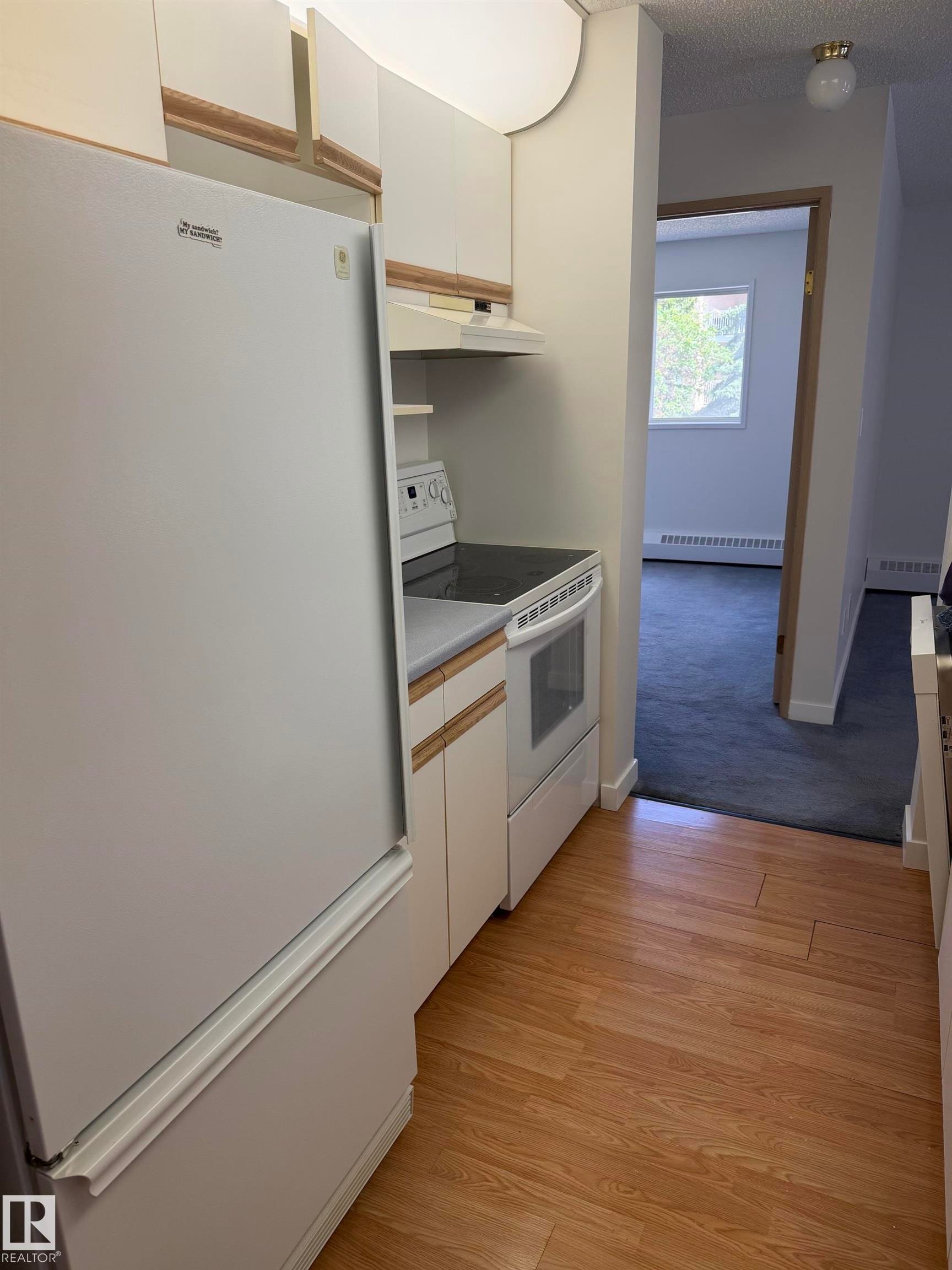 Galley kitchen with wood-finish flooring, white cabinetry, and white appliances including a refrigerator and electric range with an overhead range hood - 213 17467 98A Avenue, Edmonton, AB