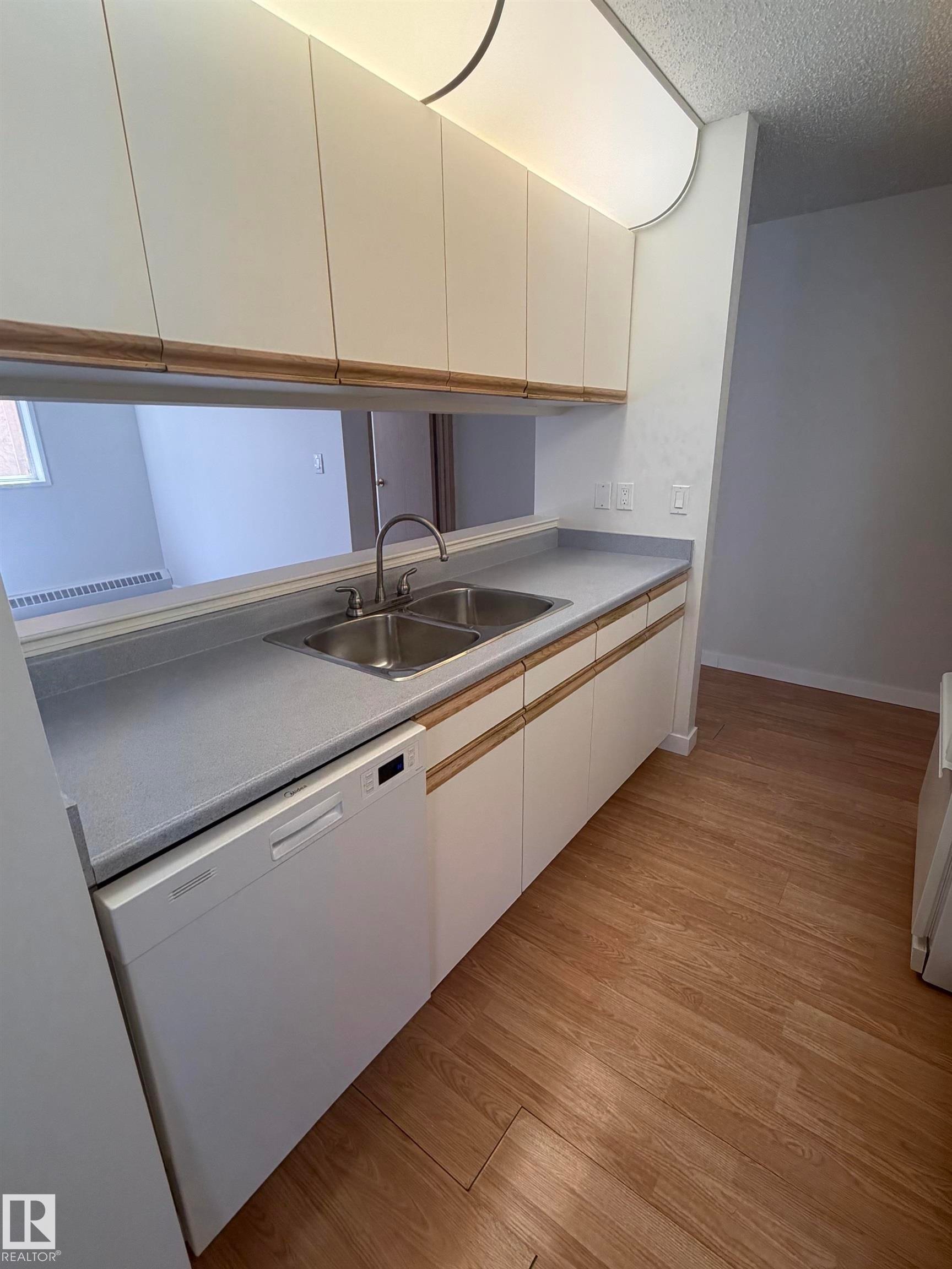 Kitchen area featuring a double basin stainless steel sink, a white built-in dishwasher, light wood-finish flooring, white cabinetry with wood trim, and a pass-through opening - 213 17467 98A Avenue, Edmonton, AB