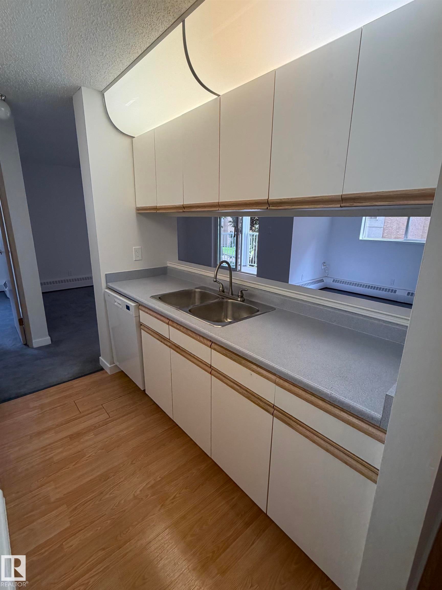Kitchenette featuring white cabinetry, a double basin stainless steel sink with a gooseneck faucet, light-toned countertops, and wood-finish flooring - 213 17467 98A Avenue, Edmonton, AB