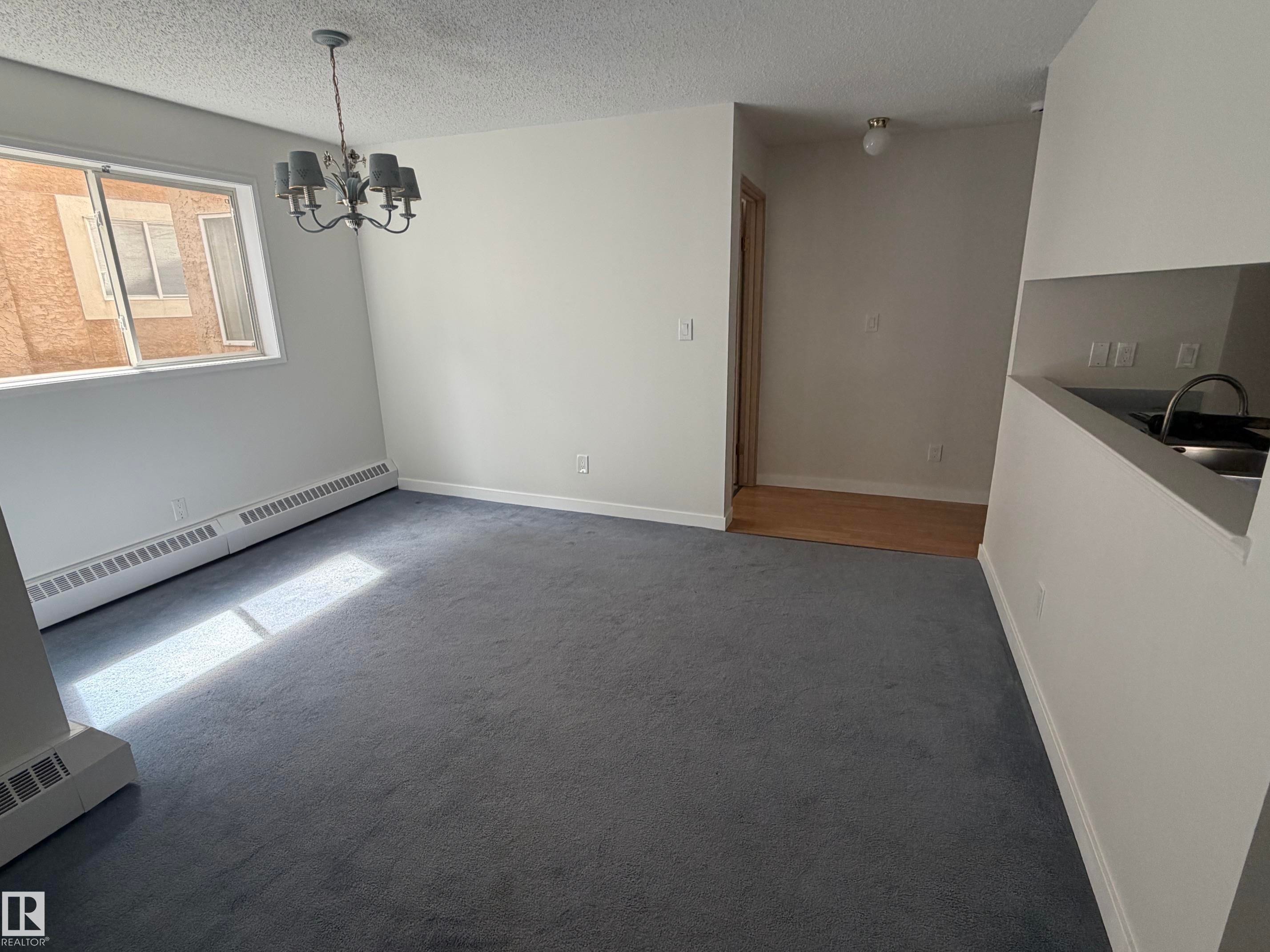 Spacious room featuring gray carpeting, a multi-light chandelier, a large window, and white baseboard trim - 213 17467 98A Avenue, Edmonton, AB