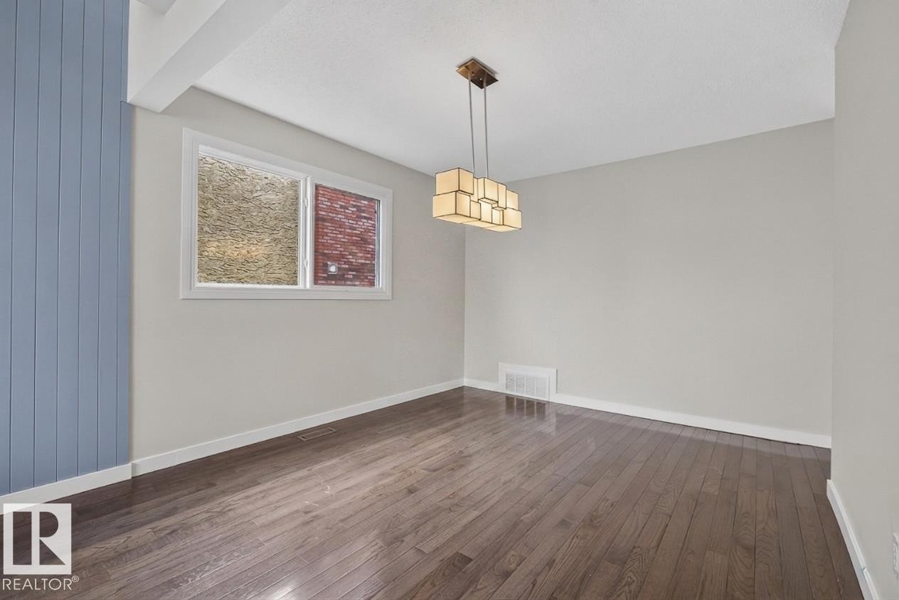Spacious interior room featuring wood-finish flooring, a modern geometric chandelier, a window with white trim, and a prominent blue vertical plank accent wall - 2712 104 Street, Edmonton, AB