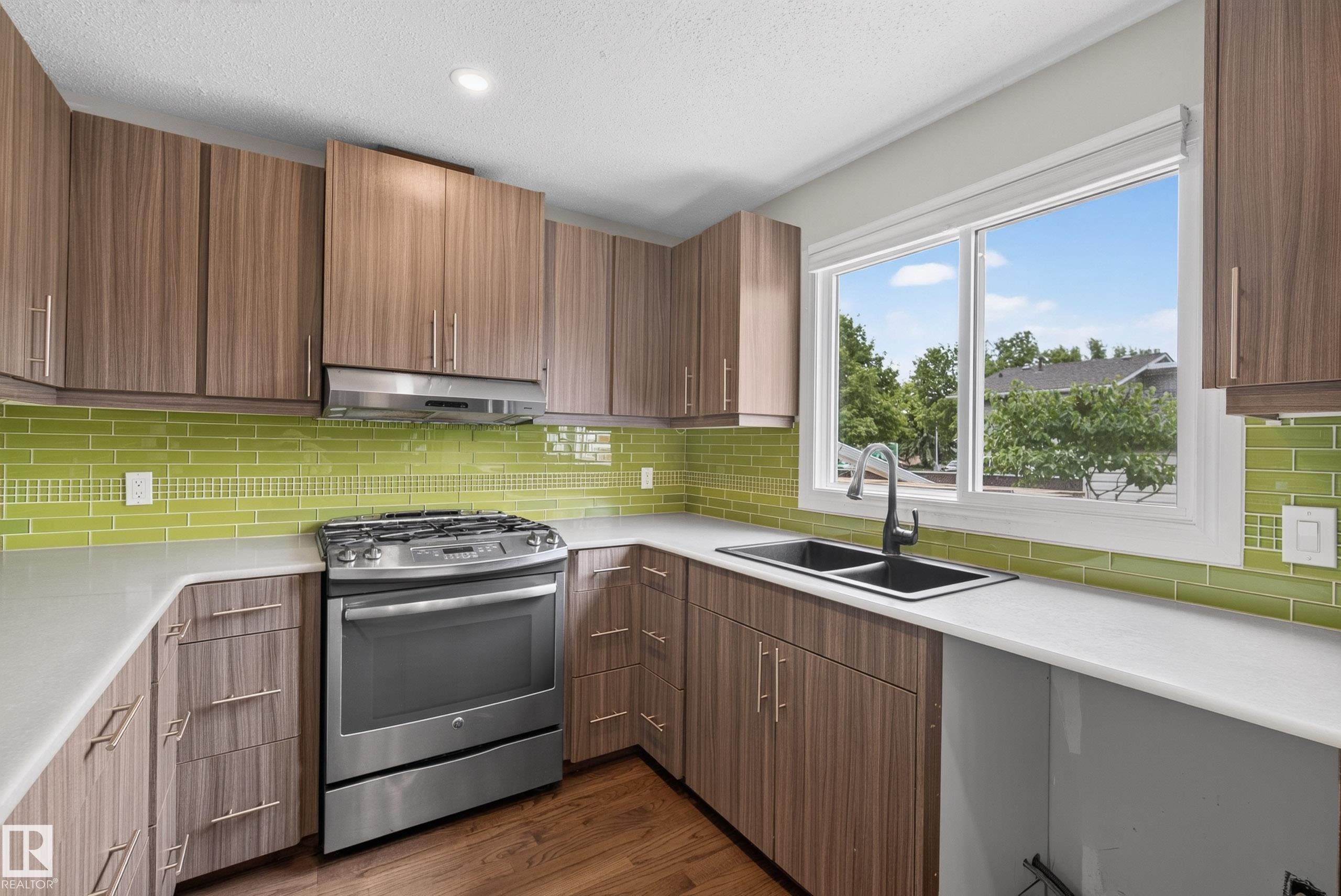 Contemporary kitchen featuring a stainless steel gas range, wood-grain cabinetry, light-toned countertops, a vibrant green subway tile backsplash, and a double basin sink with a matte black faucet - 2712 104 Street, Edmonton, AB