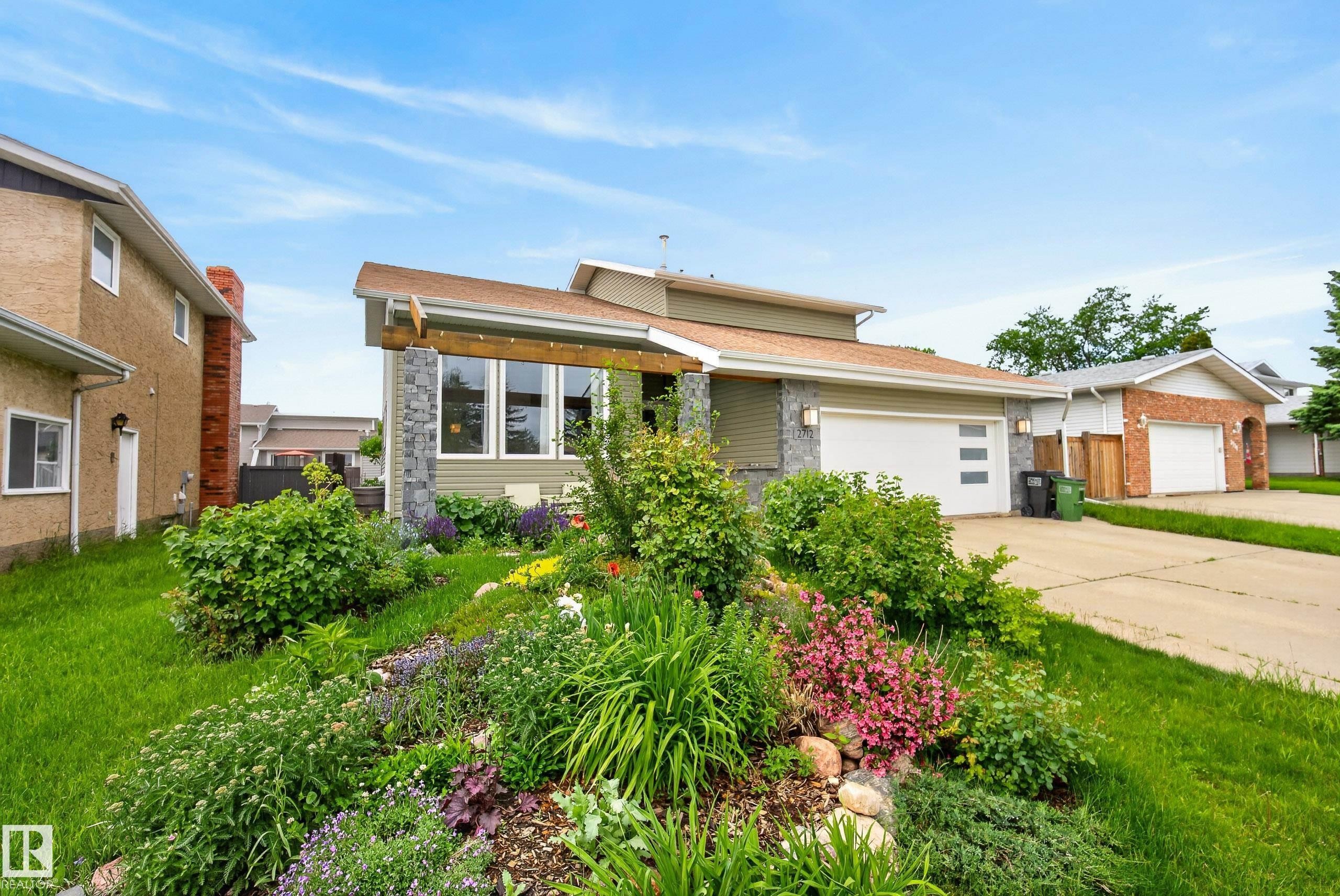 Modern split-level residence featuring a multi-pane garage door, stone and siding exterior, and a sloped front yard with established landscaping and various plantings - 2712 104 Street, Edmonton, AB