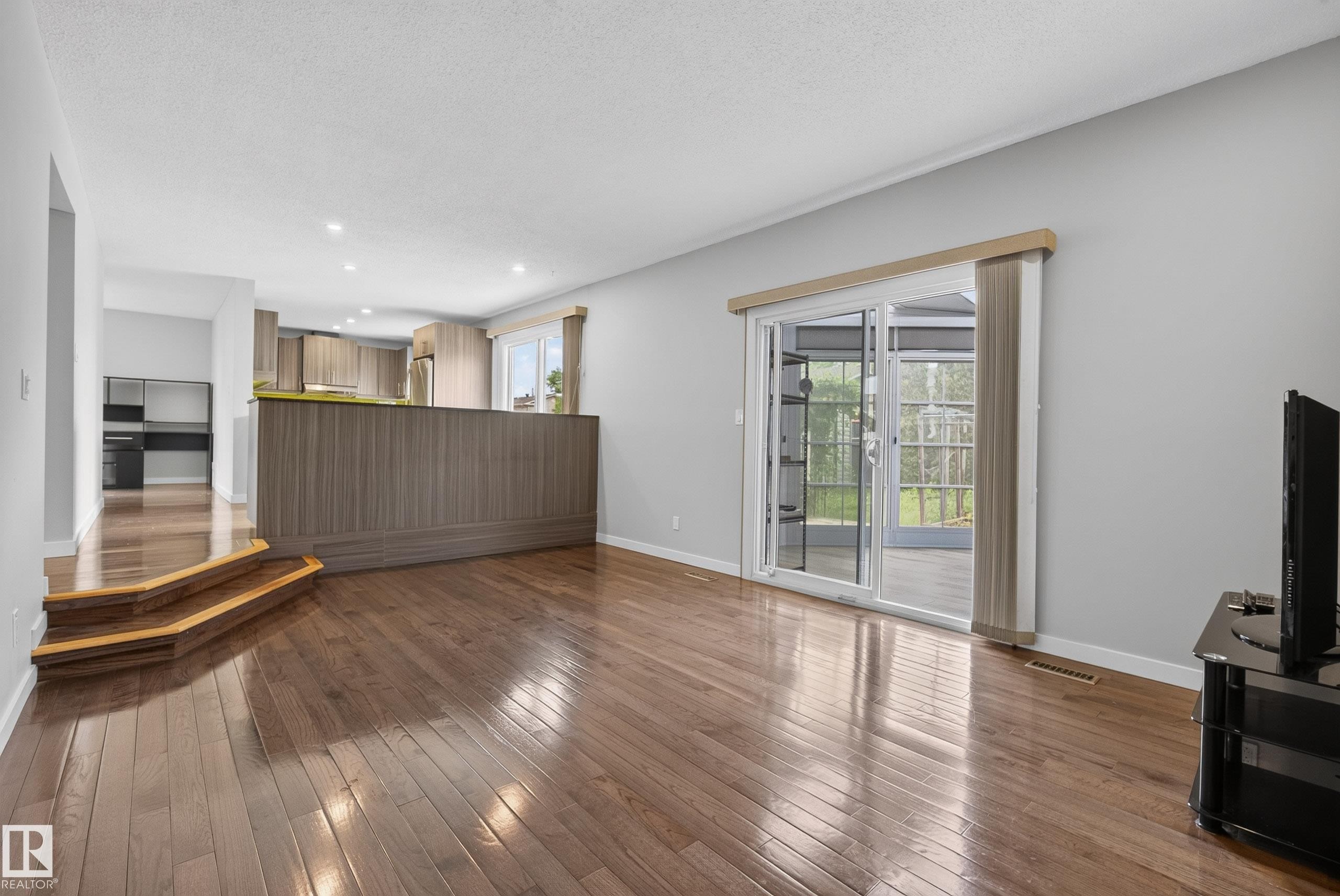 Open-concept living area featuring rich wood-finish flooring, a contemporary wood-paneled breakfast bar, a modern kitchen with stainless steel appliances, and sliding glass doors leading to an enclosed patio - 2712 104 Street, Edmonton, AB