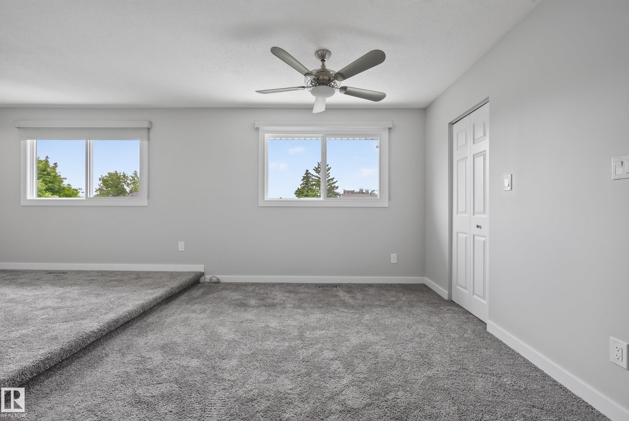 Spacious room featuring neutral carpeting, light gray walls, a ceiling fan with integrated lighting, two windows with white trim, and a bi-fold closet door - 2712 104 Street, Edmonton, AB
