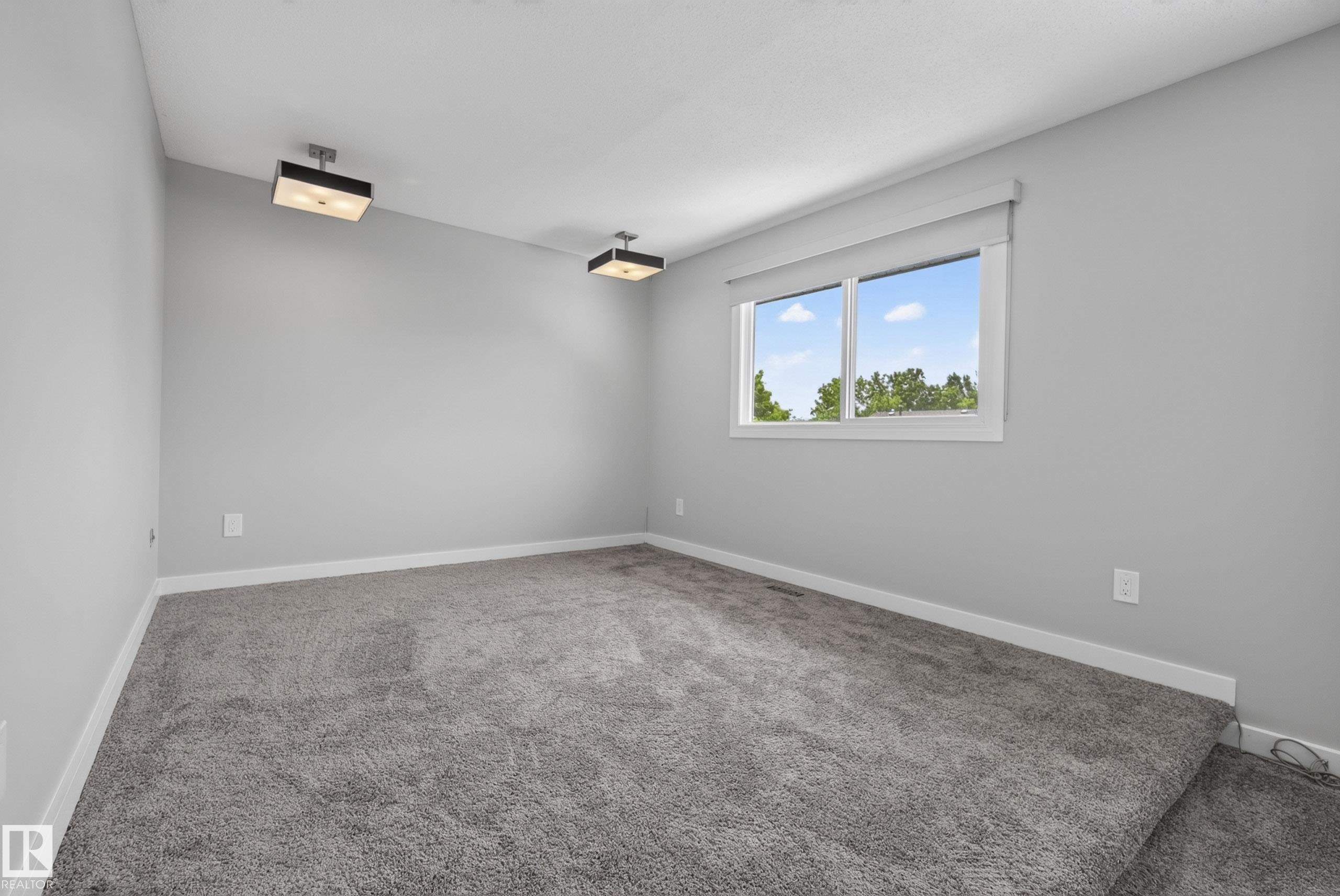 Spacious room featuring light gray walls, gray carpeting, a large window, and two ceiling-mounted light fixtures - 2712 104 Street, Edmonton, AB