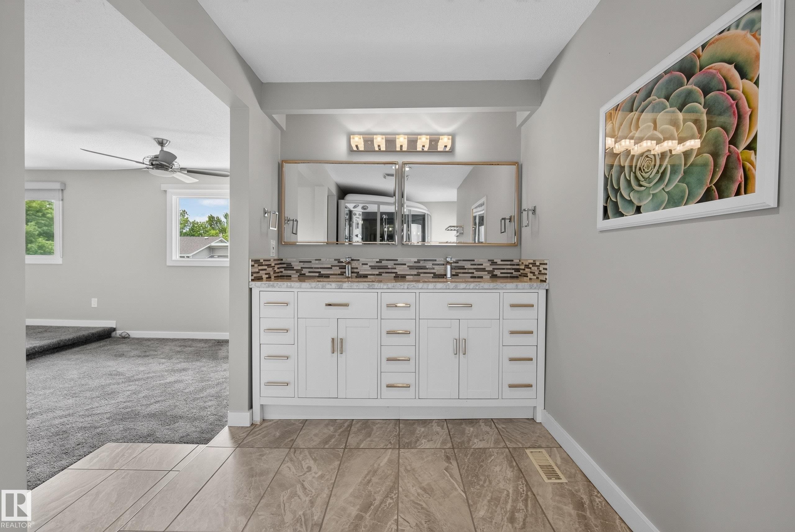 Bathroom vanity featuring dual rectangular sinks, a stone countertop, white shaker-style cabinetry with gold-tone hardware, and a mosaic tile backsplash - 2712 104 Street, Edmonton, AB