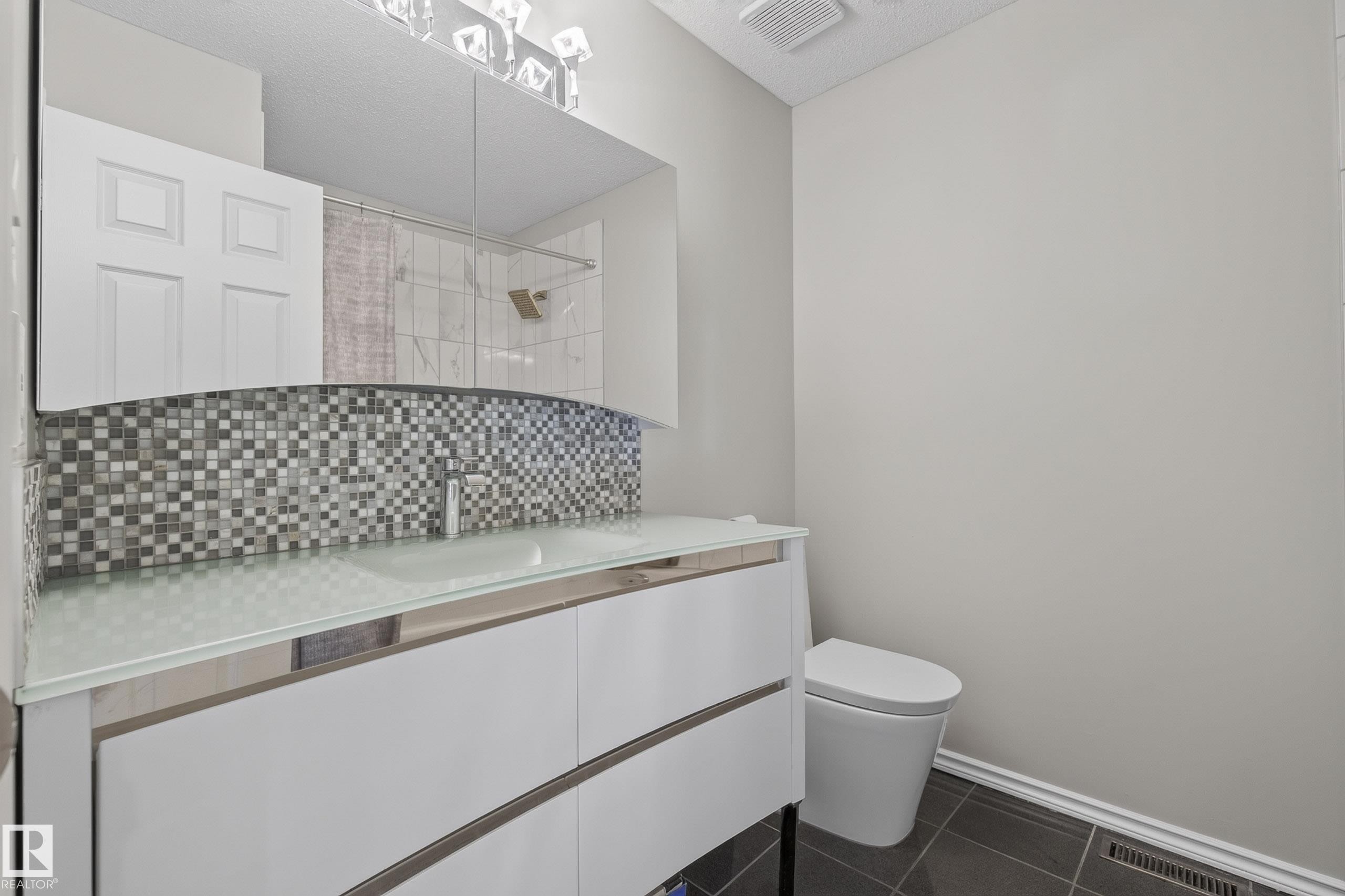 Bathroom vanity featuring a frosted glass countertop, contemporary white cabinetry, and a mosaic tile backsplash - 2712 104 Street, Edmonton, AB