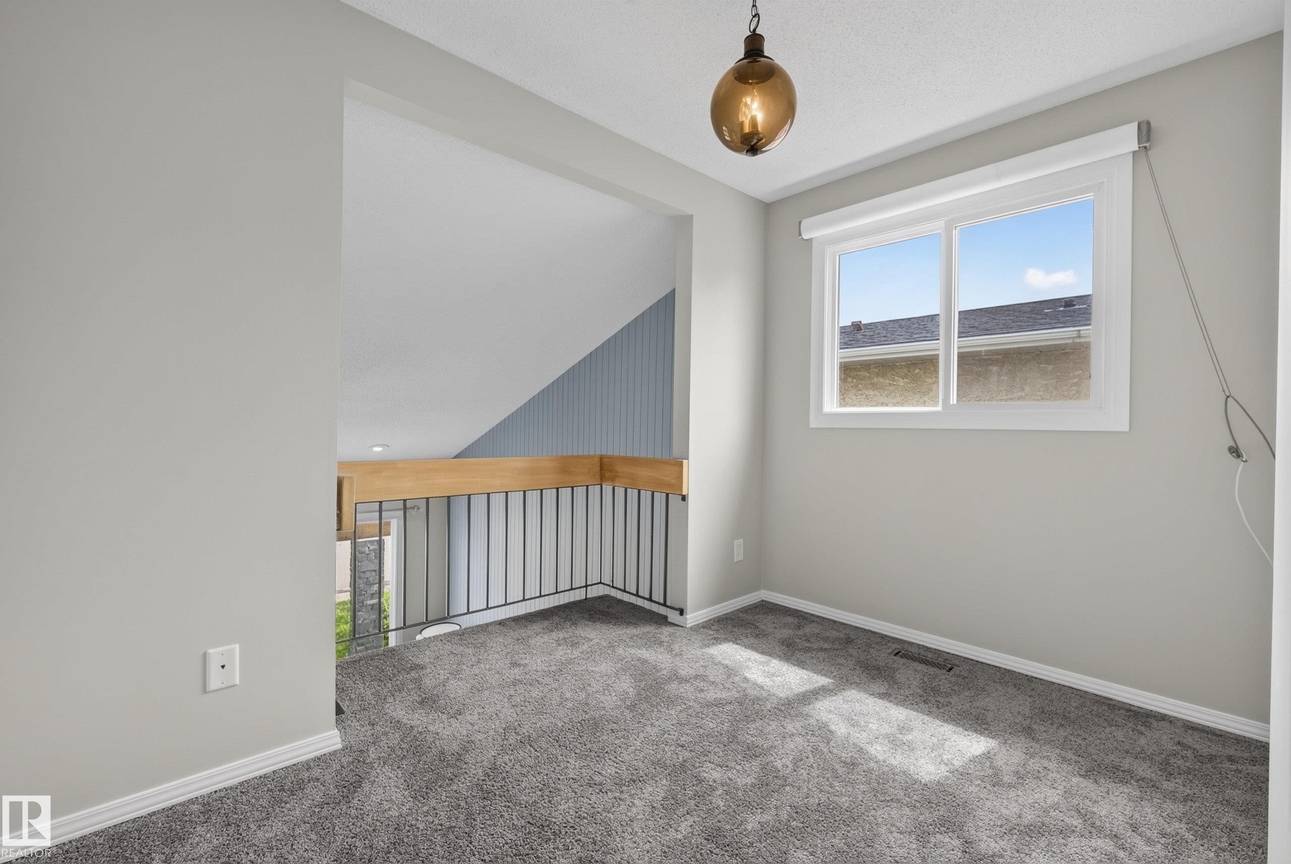 Carpeted open-concept space featuring a large window with roller shade, a decorative pendant light fixture, a wood-finish railing with black metal spindles, and light gray painted walls - 2712 104 Street, Edmonton, AB