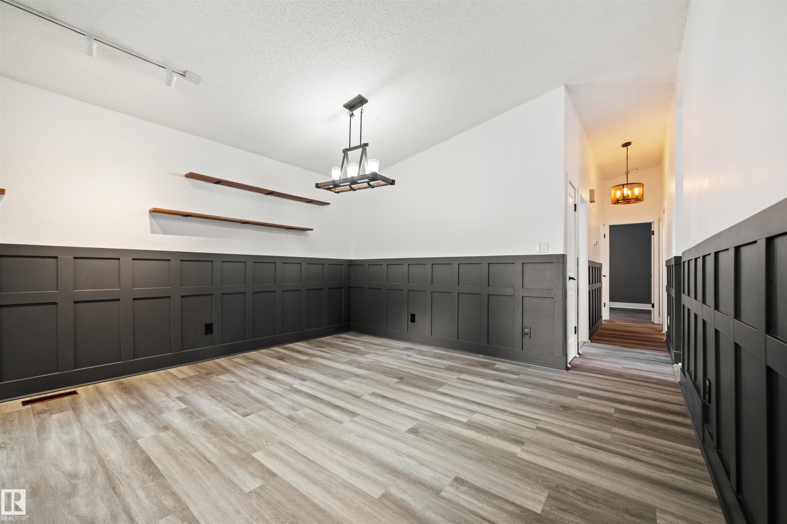 Spacious interior featuring contemporary wainscoting, wood-finish flooring, a modern chandelier, track lighting, and floating wood shelves - 28 Goodridge Drive, St. Albert, AB