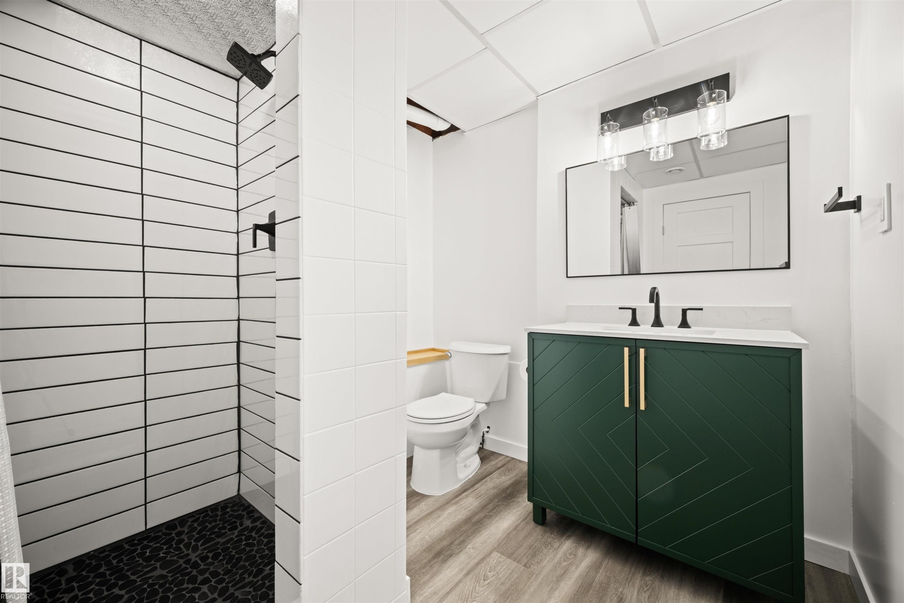 Contemporary bathroom featuring a dark green vanity with brass hardware, a rectangular vanity mirror, a three-light fixture, and a tiled shower with black pebble flooring and a matte black shower head - 28 Goodridge Drive, St. Albert, AB