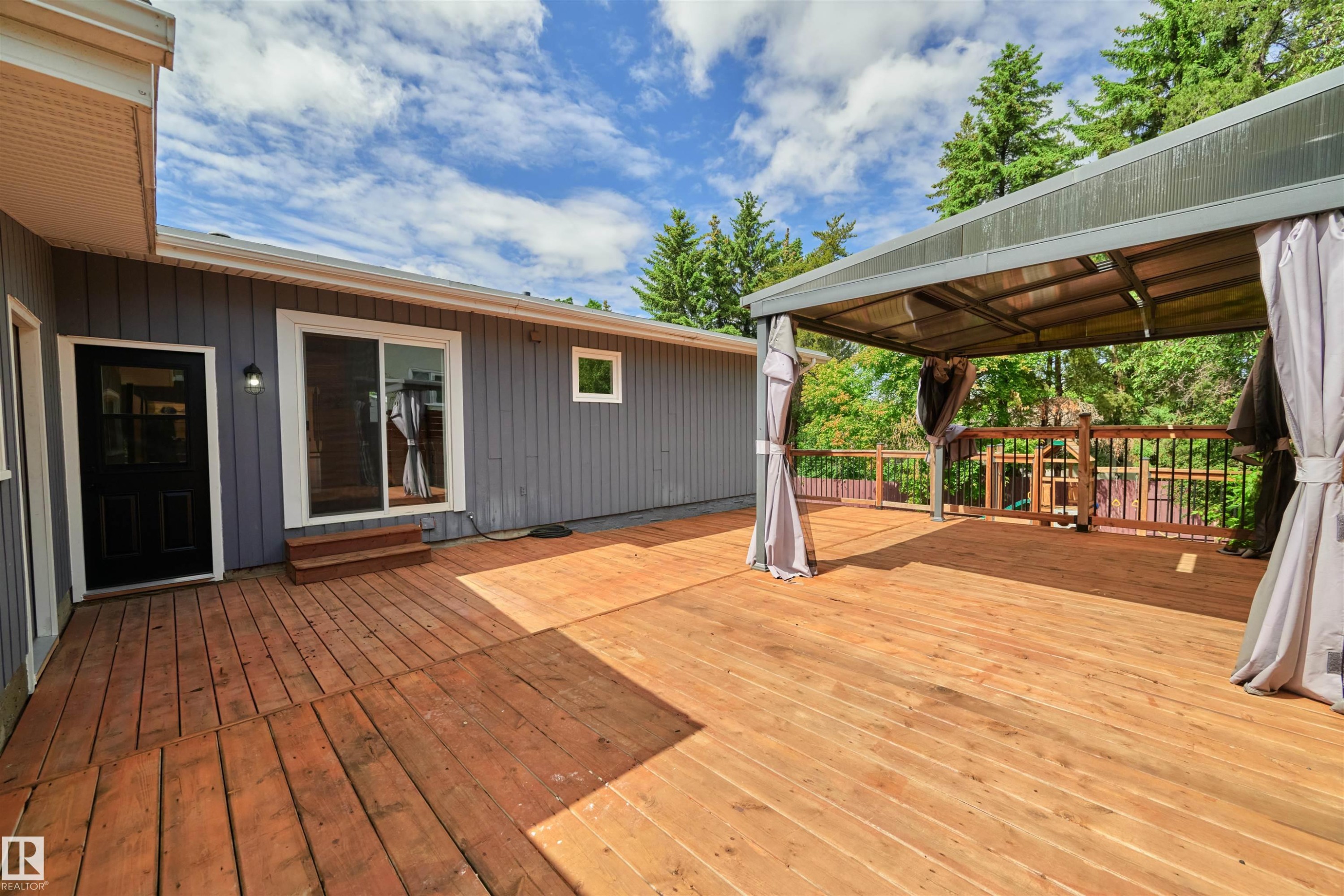 Expansive wood-finish deck featuring a covered pergola with privacy drapes, a sliding glass door entry, a separate solid black entry door, and a wooden railing - 28 Goodridge Drive, St. Albert, AB