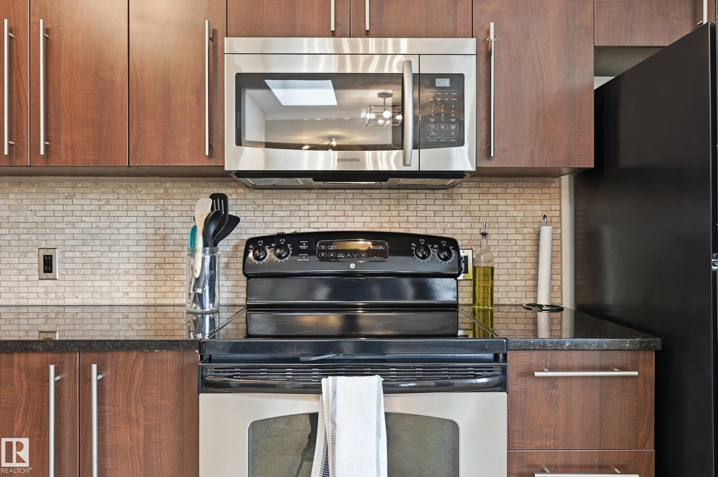 Kitchen featuring wood-finish cabinetry with brushed nickel hardware, a mosaic tile backsplash, black stone countertops, and stainless steel appliances - 12326 92 Street, Edmonton, AB - Indoor Photo Showing Kitchen