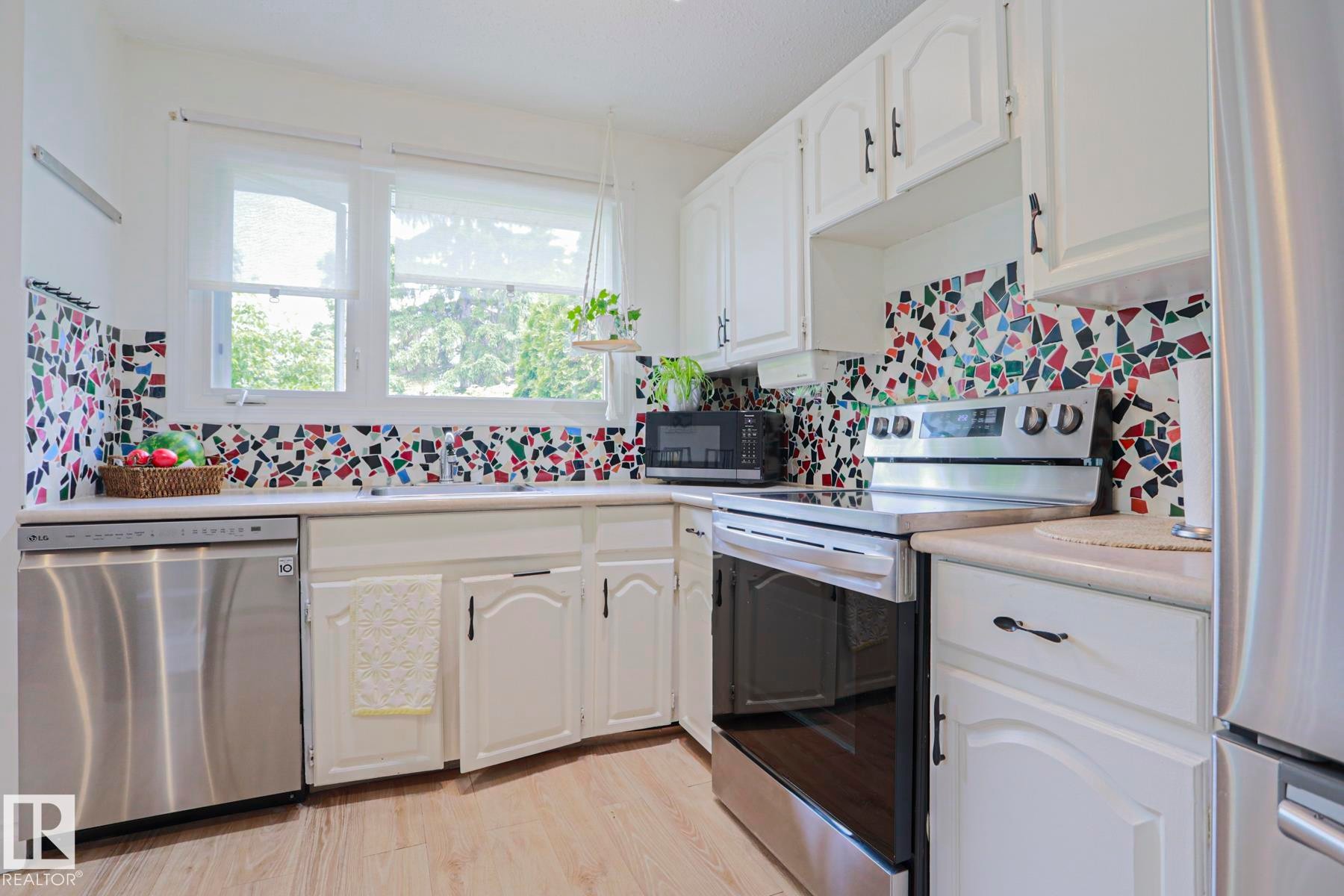Bright kitchen featuring white cabinetry, a colorful mosaic tile backsplash, stainless steel dishwasher, and a freestanding range with a flat cooktop - 2007 67 Street, Edmonton, AB