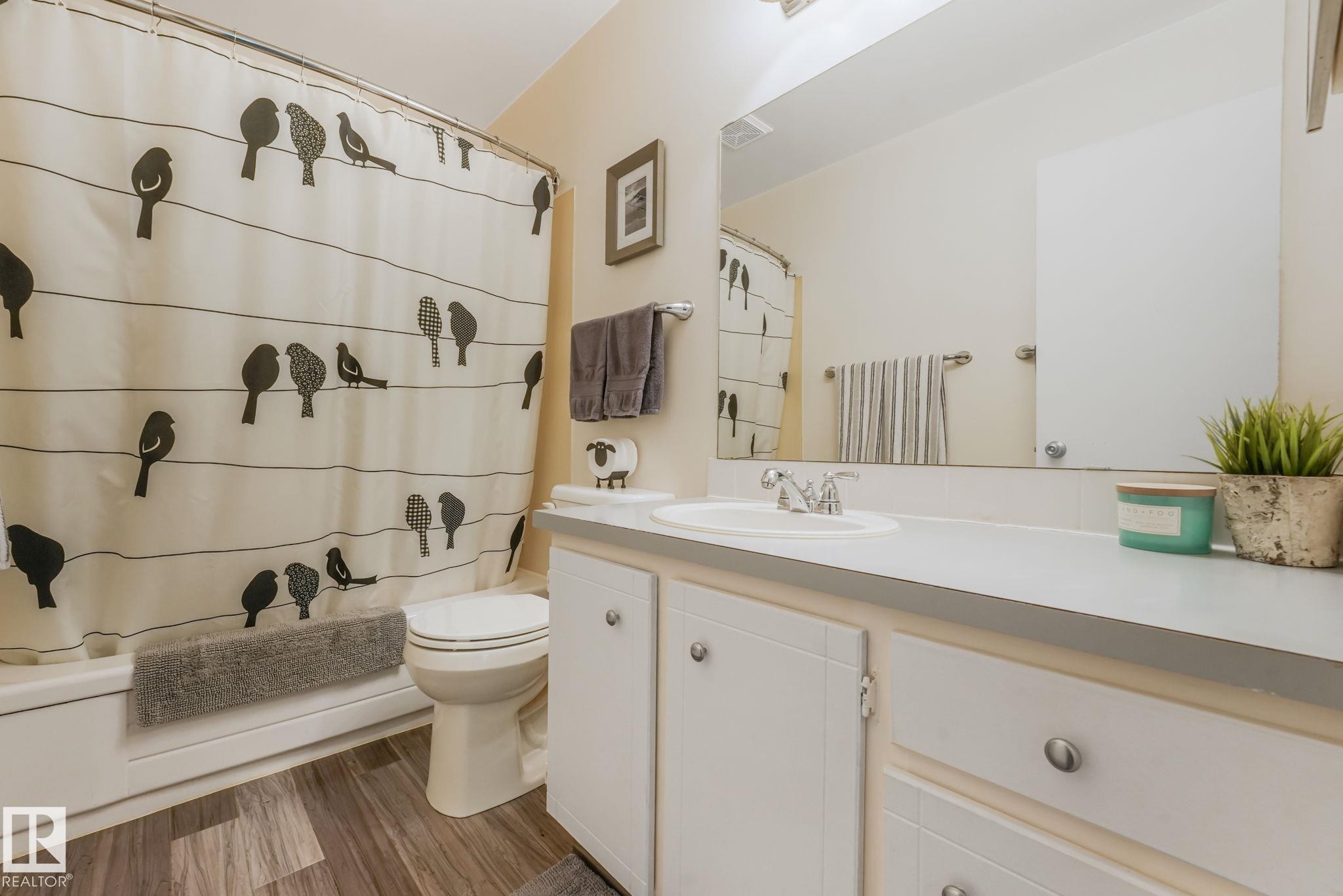 Bathroom featuring a white vanity with integrated sink, a full-width mirror, a white bathtub with a shower, and wood-finish flooring - 17237 104 Street, Edmonton, AB