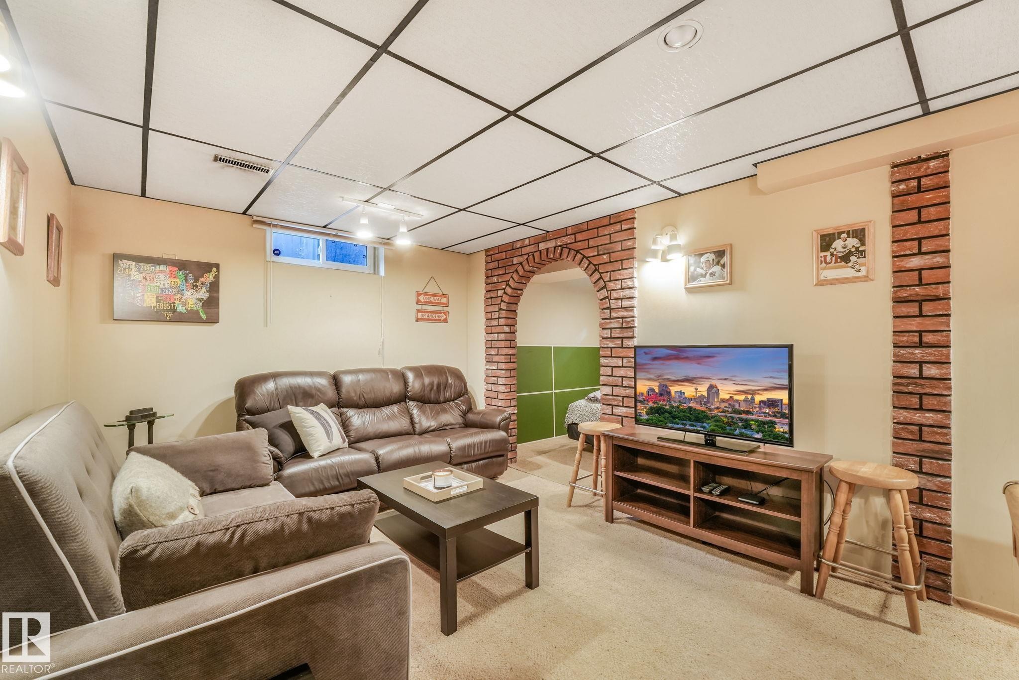 Finished lower level featuring an exposed grid ceiling, carpeted flooring, a brick archway, a window, and wall sconces - 17237 104 Street, Edmonton, AB