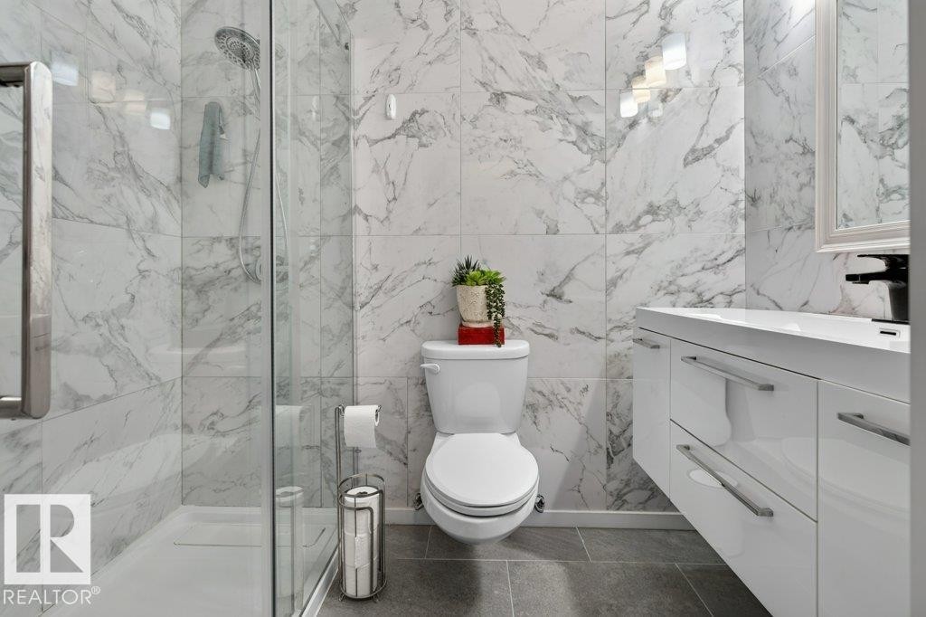 Contemporary bathroom featuring large format marble-look wall tiles, a frameless glass shower enclosure, a white floating vanity with integrated sink, and dark gray tile flooring - 6916 92A Avenue, Edmonton, AB