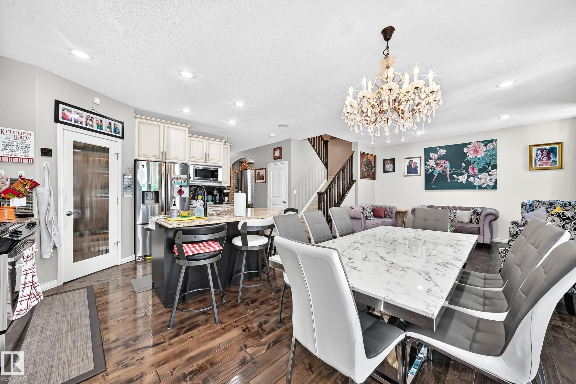 Open-concept living space featuring wood-finish flooring, a kitchen island with seating, stainless steel appliances, a crystal chandelier, and a staircase with dark wood railings - 12410 171A Avenue, Edmonton, AB