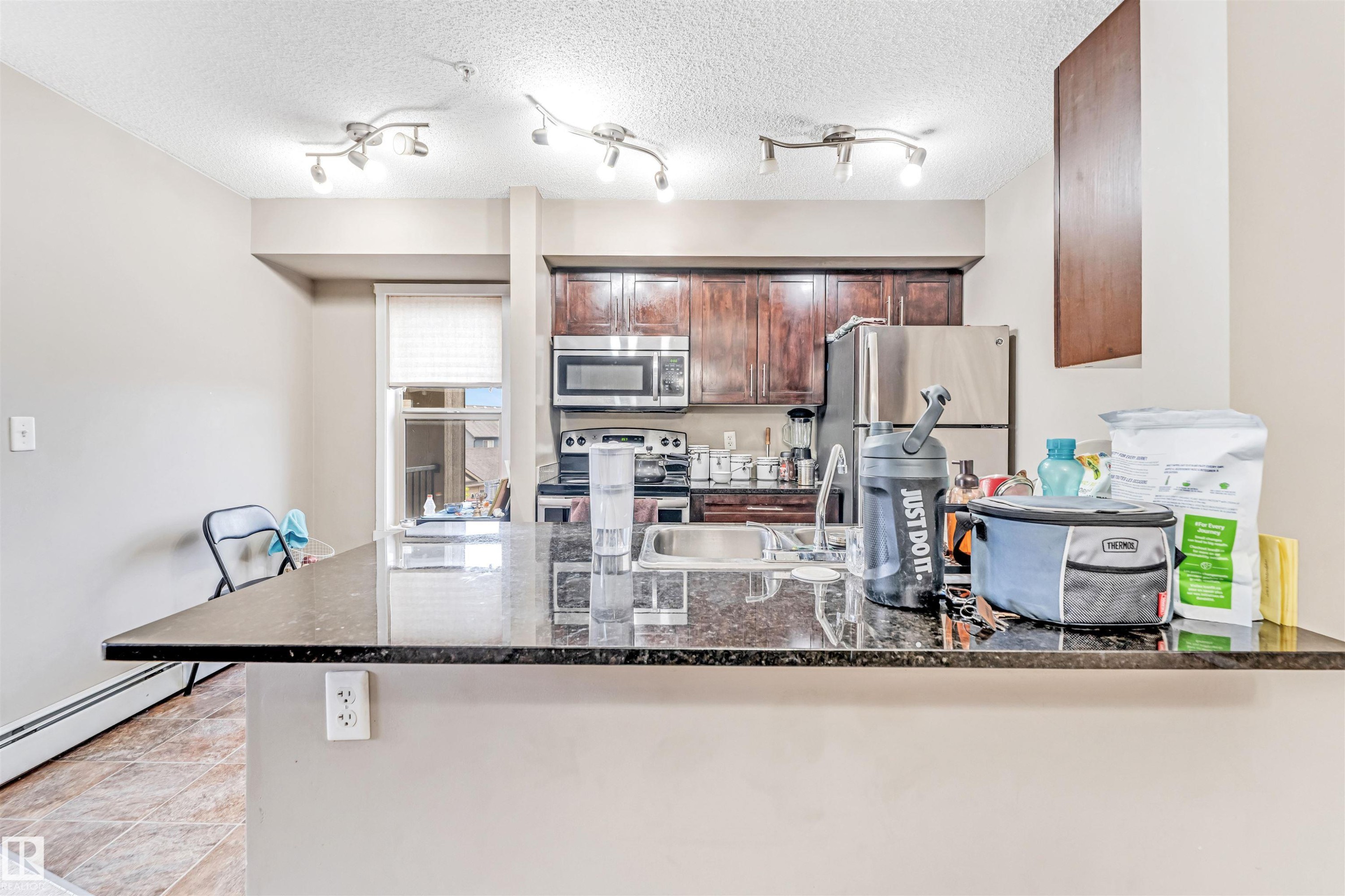 Kitchen featuring an expansive stone-finish countertop with an undermount sink, dark wood-finish cabinetry, and stainless steel appliances including a refrigerator and microwave - 225 111 Watt Common, Edmonton, AB