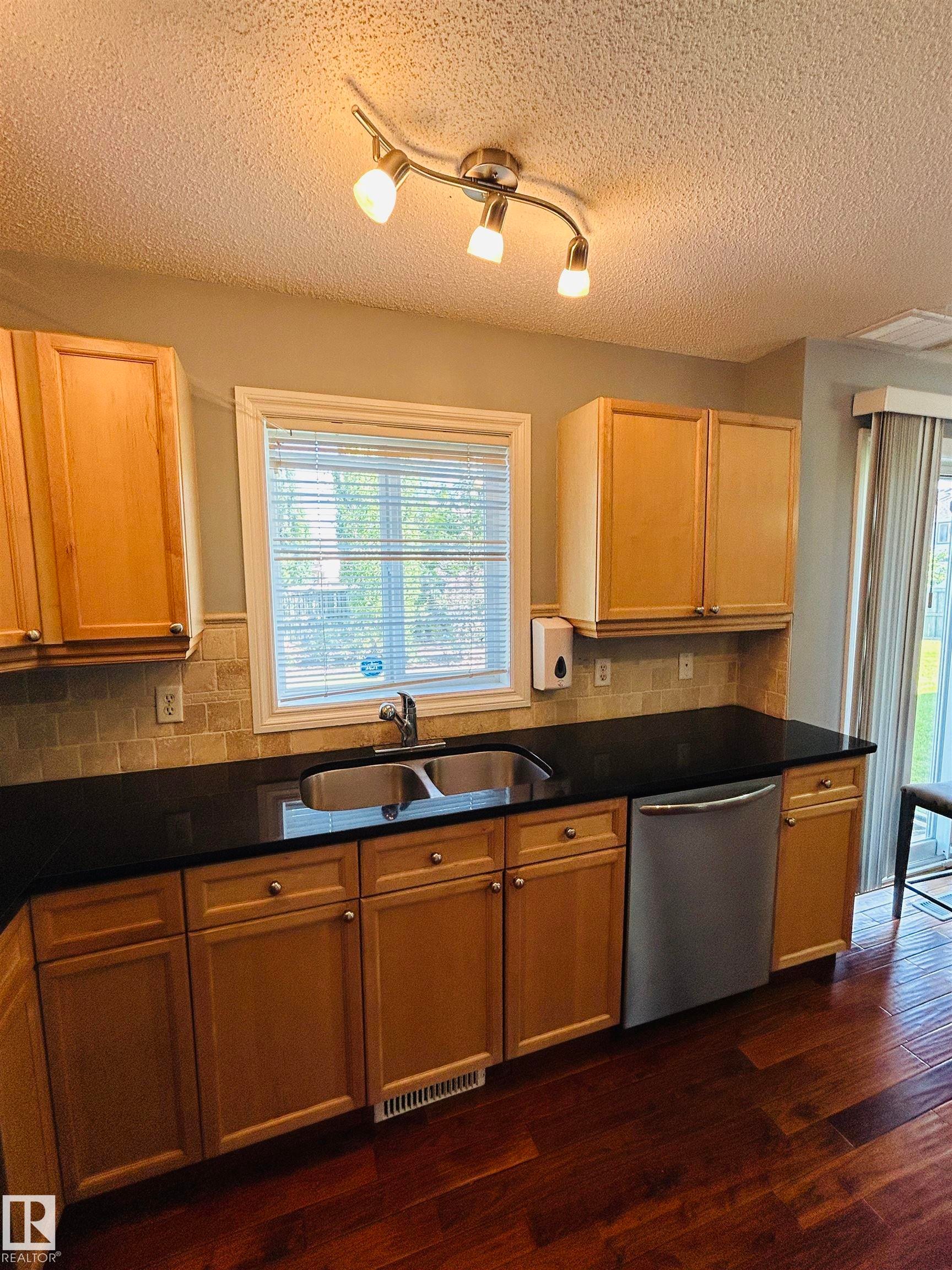 Kitchen featuring wood-finish flooring, light wood cabinetry, black countertops, and a stainless steel double basin sink with a matching faucet - 89 12050 17 Avenue, Edmonton, AB