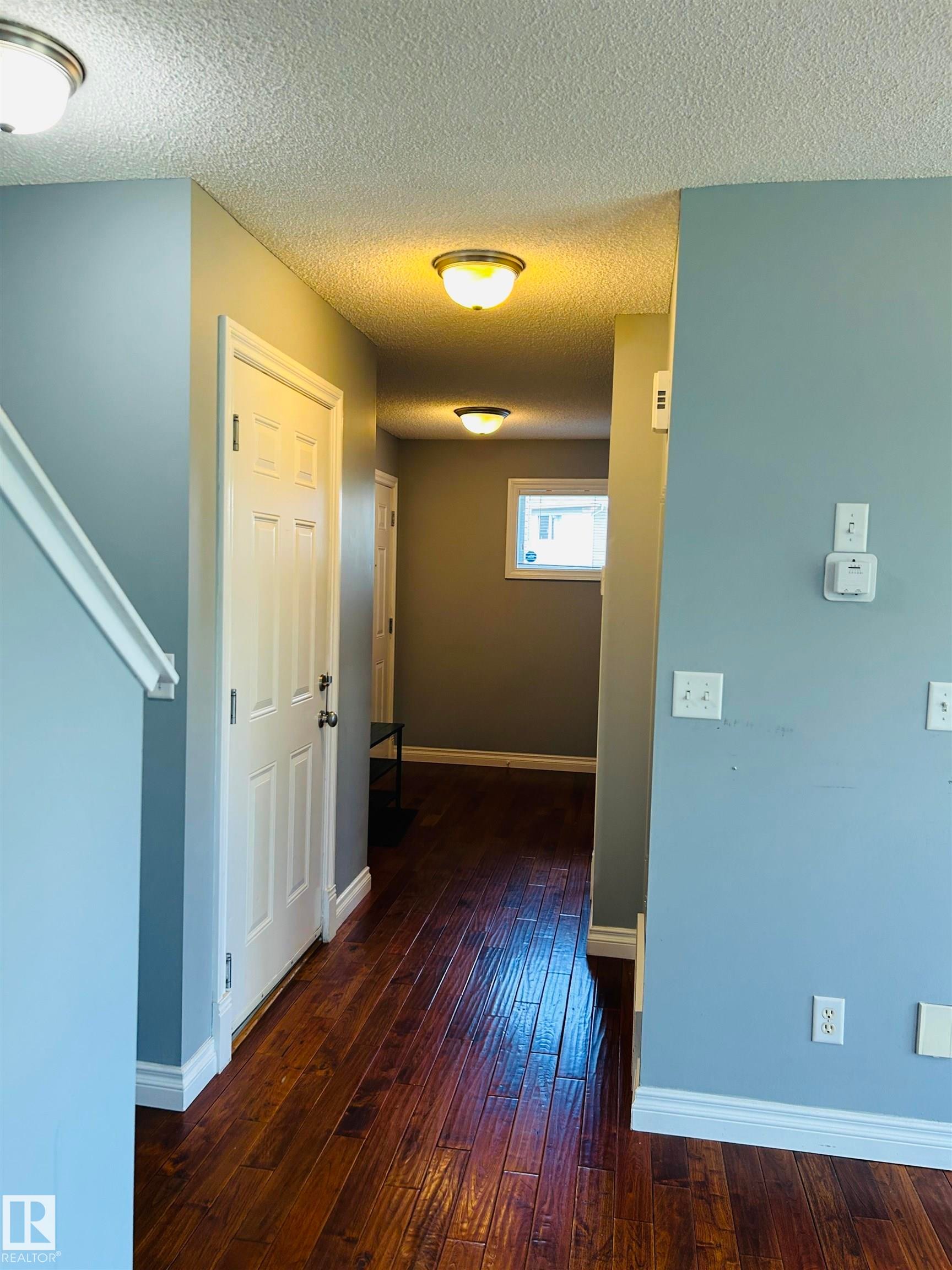 Hallway featuring rich wood-finish flooring, light blue wall paint, and multiple flush-mount ceiling fixtures - 89 12050 17 Avenue, Edmonton, AB