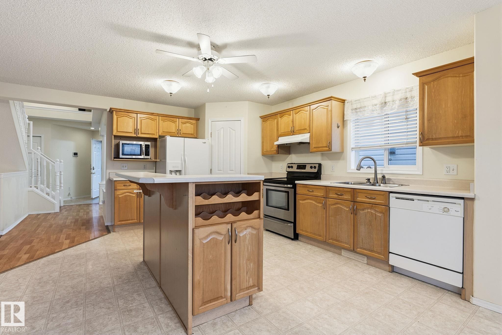 Kitchen featuring wood cabinetry, a center island with built-in storage and wine rack, stainless steel appliances, and a double basin sink - 1906 37B Av Nw, Edmonton, AB - Indoor Photo Showing Kitchen With Double Sink
