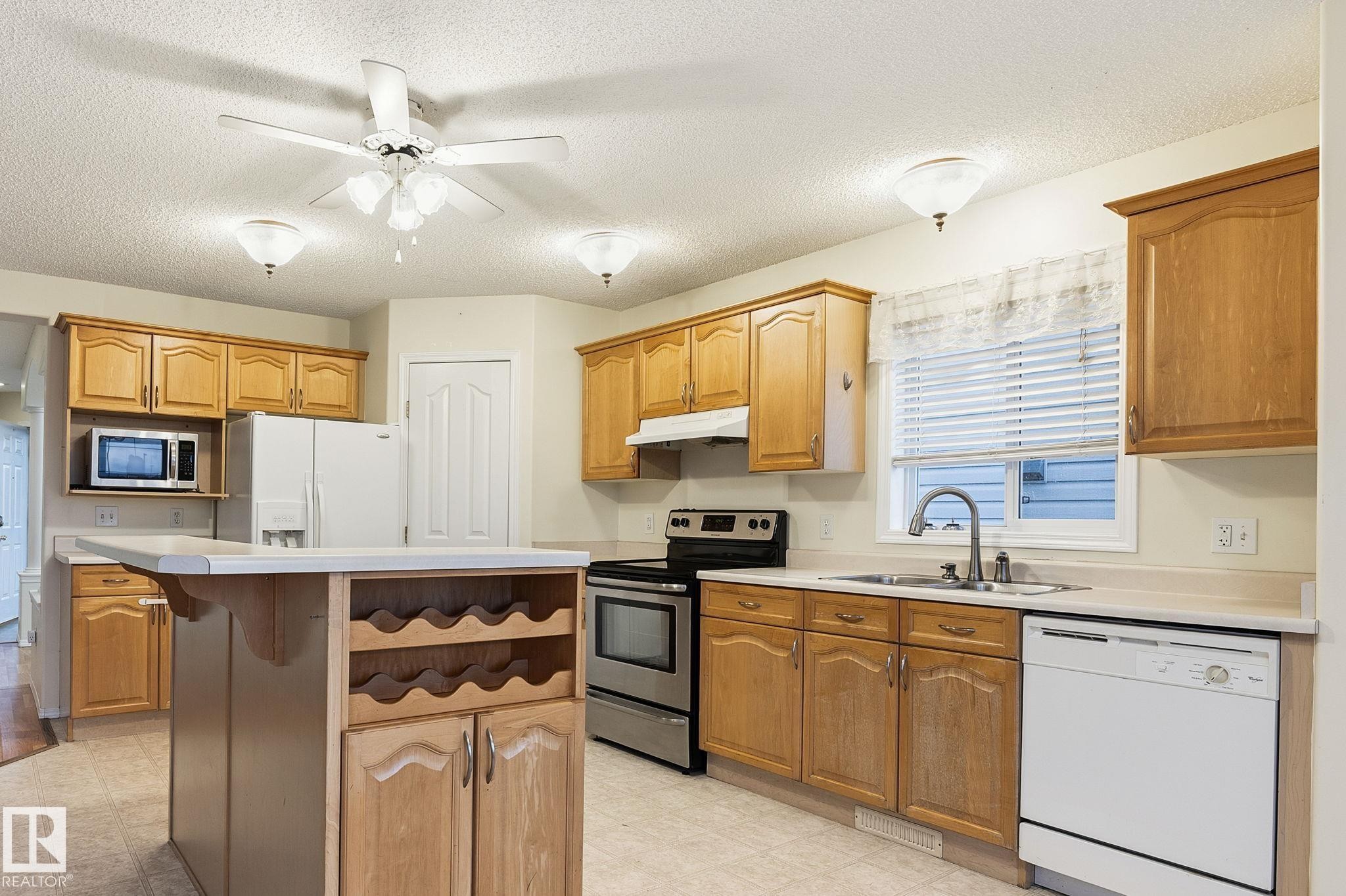 Kitchen featuring wood-finish cabinetry, a central island with integrated wine storage, and a double basin sink with a chrome faucet - 1906 37B Av Nw, Edmonton, AB - Indoor Photo Showing Kitchen With Double Sink