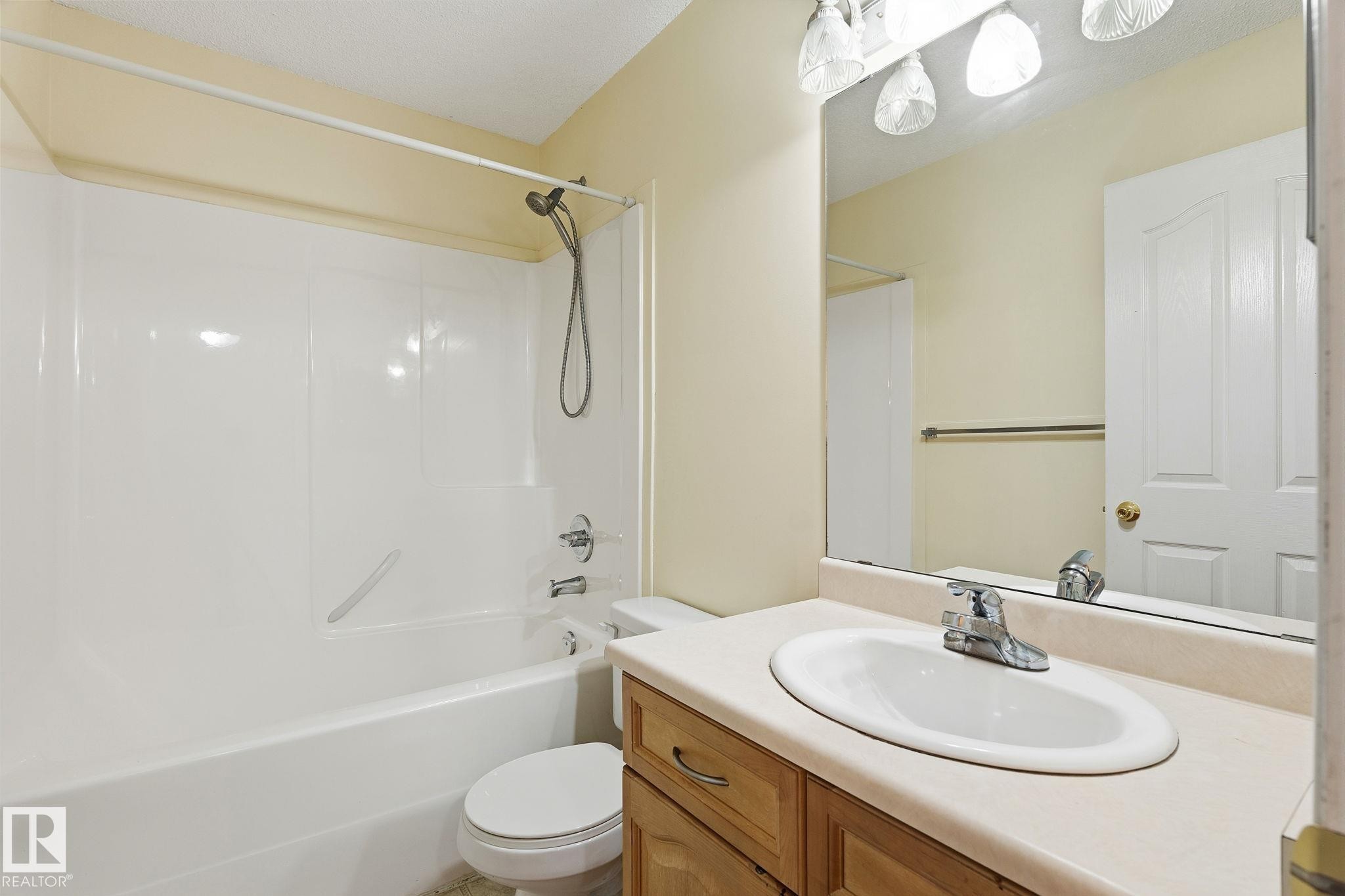 Bathroom featuring a combination bathtub and shower with white surround, a single vanity with an integrated sink, a large mirror, and a three-light fixture - 1906 37B Av Nw, Edmonton, AB - Indoor Photo Showing Bathroom