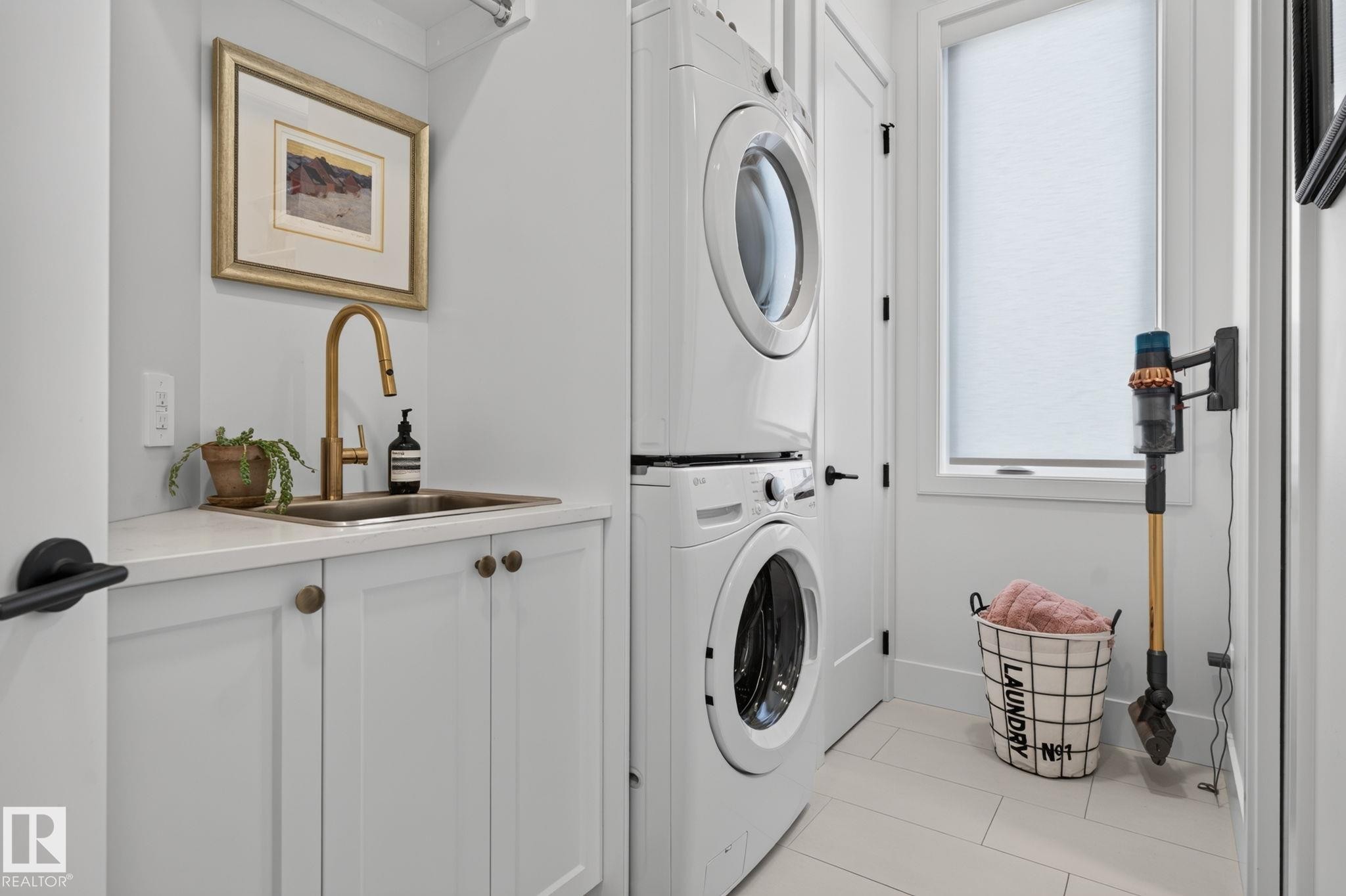 Dedicated laundry space featuring a stacked washer and dryer, white shaker-style cabinetry, a built-in sink with a gooseneck faucet, light-toned tile flooring, and a window for natural illumination - 14 Jane Point(E), St. Albert, AB